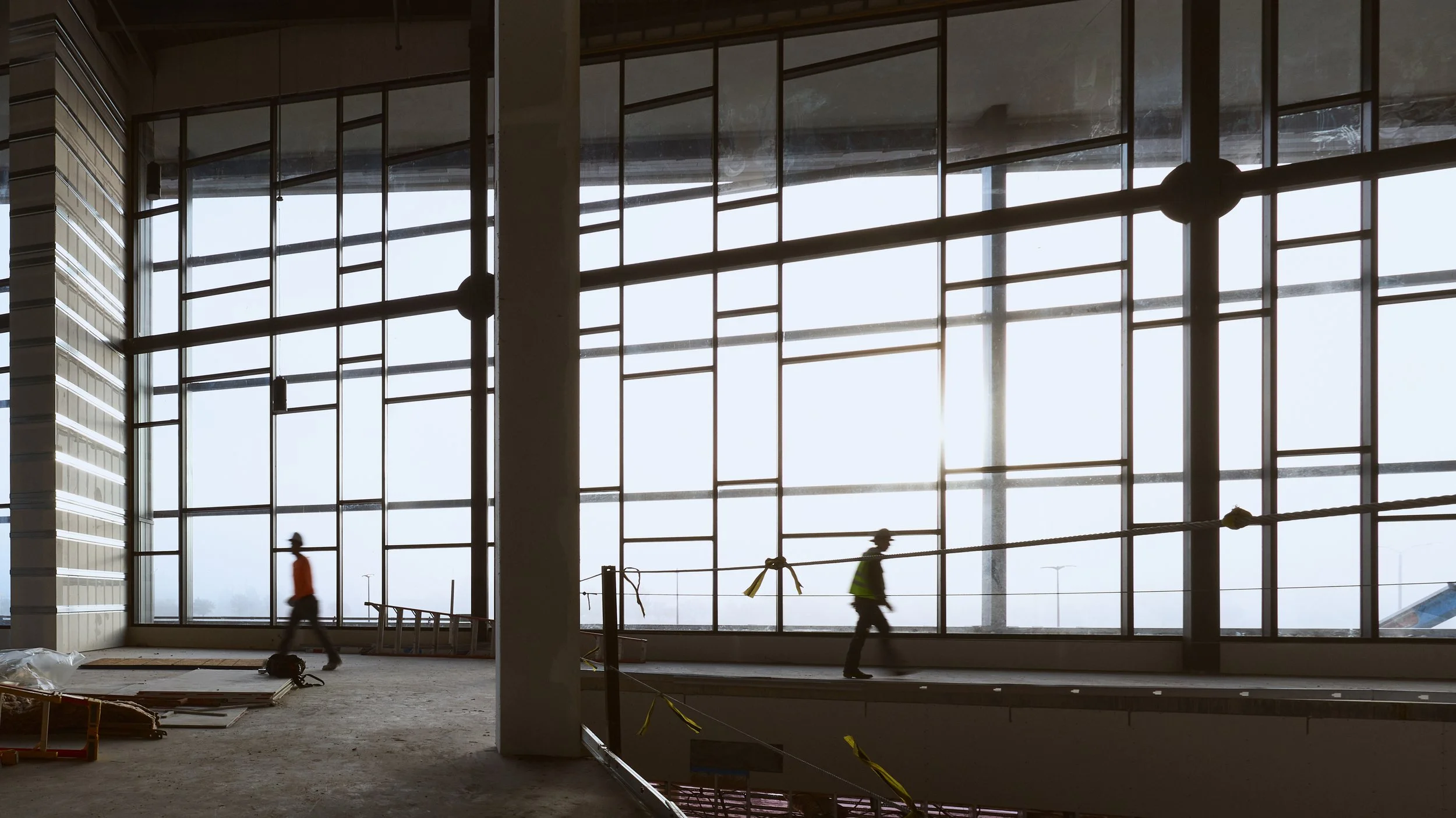 Interior of an airport terminal under construction, with large glass windows allowing bright sunlight to enter and workers walking inside.