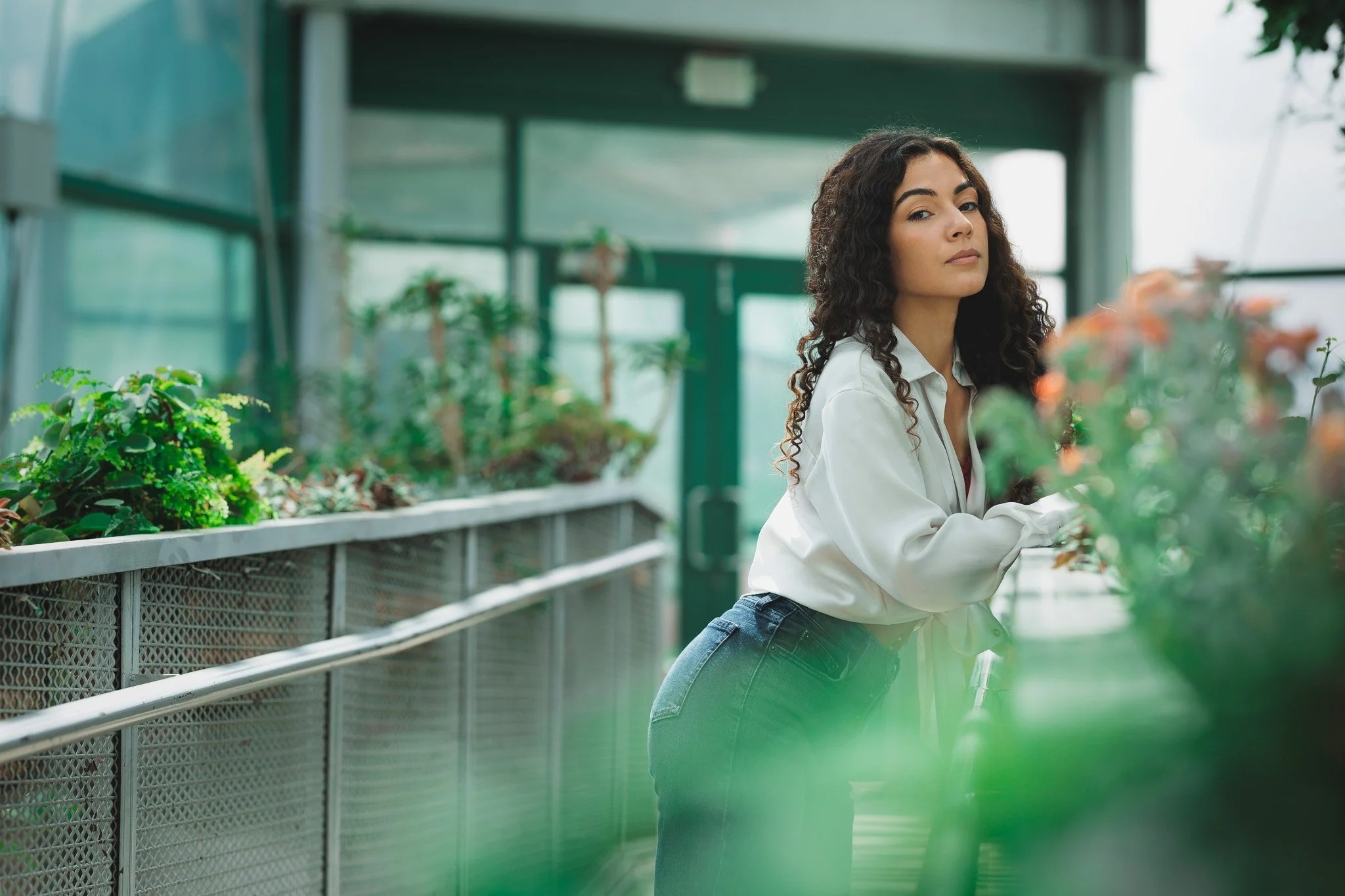 Woman with curly hair leaning on a green plant display in a greenhouse or plant shop.