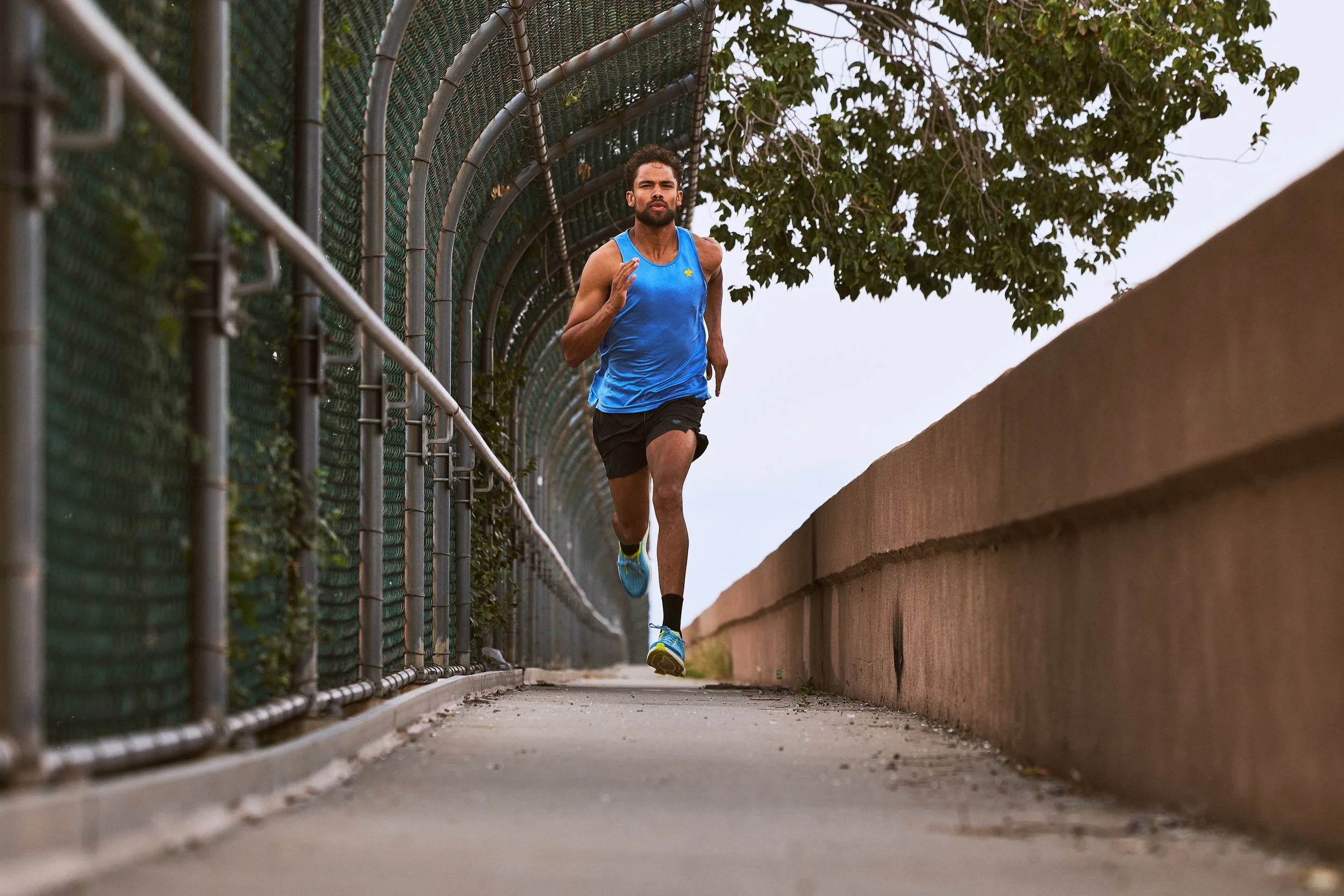 Man running outdoors on a path, seen from a low angle, with a metal fence and overhanging trees along the side.