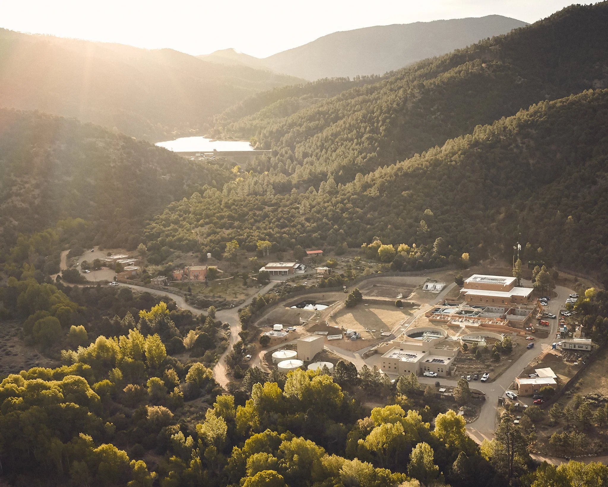 An aerial view of a hilly area with dense trees, a water reservoir, and several buildings, possibly a small community or facility, with a mountainous background and sunlight illuminating the scene.