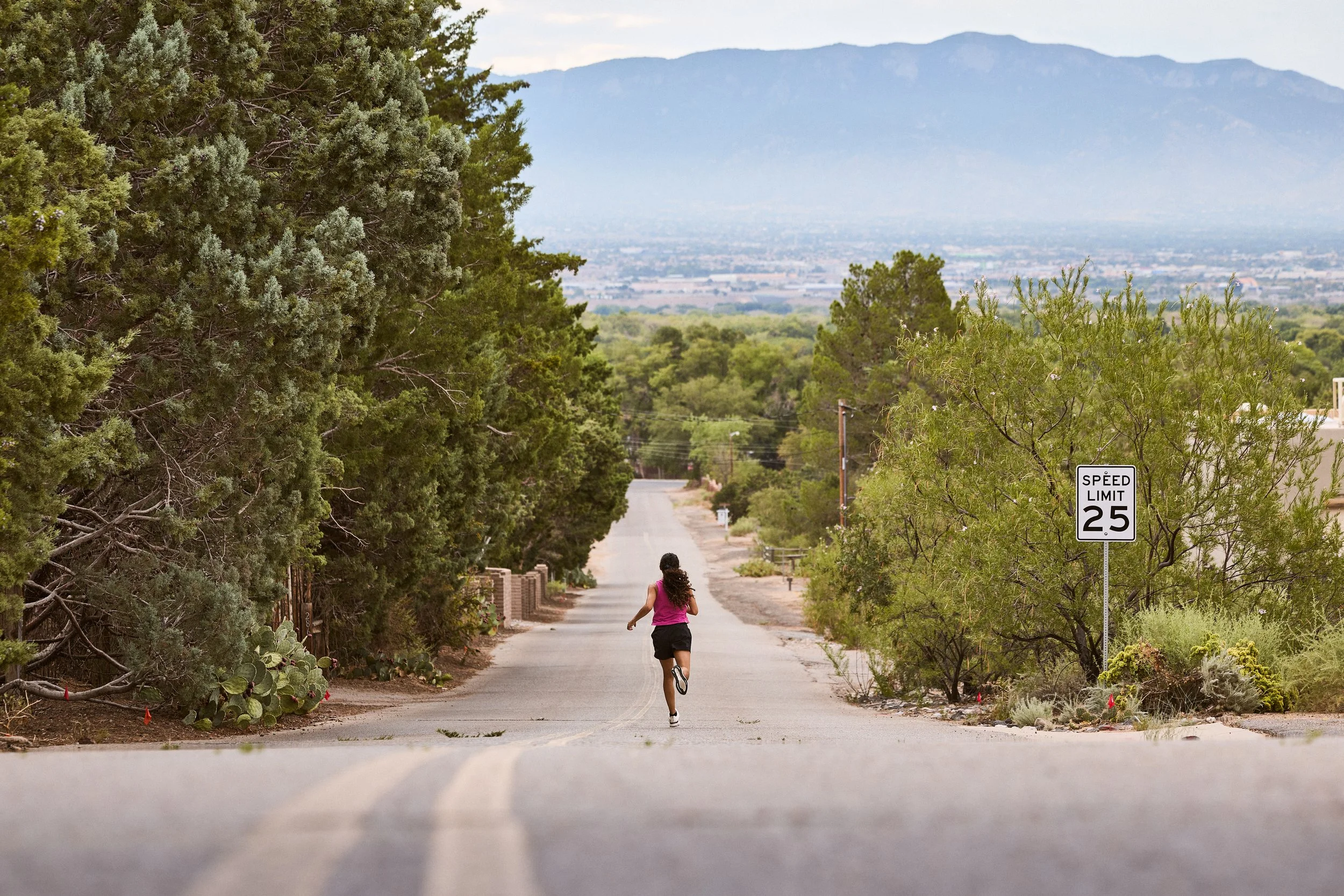 A person jogging along a quiet, paved street in a suburban or semi-rural area, lined with bushes and trees, with a mountain and a cityscape visible in the distance under a cloudy sky. A speed limit sign indicates a limit of 25 miles per hour.