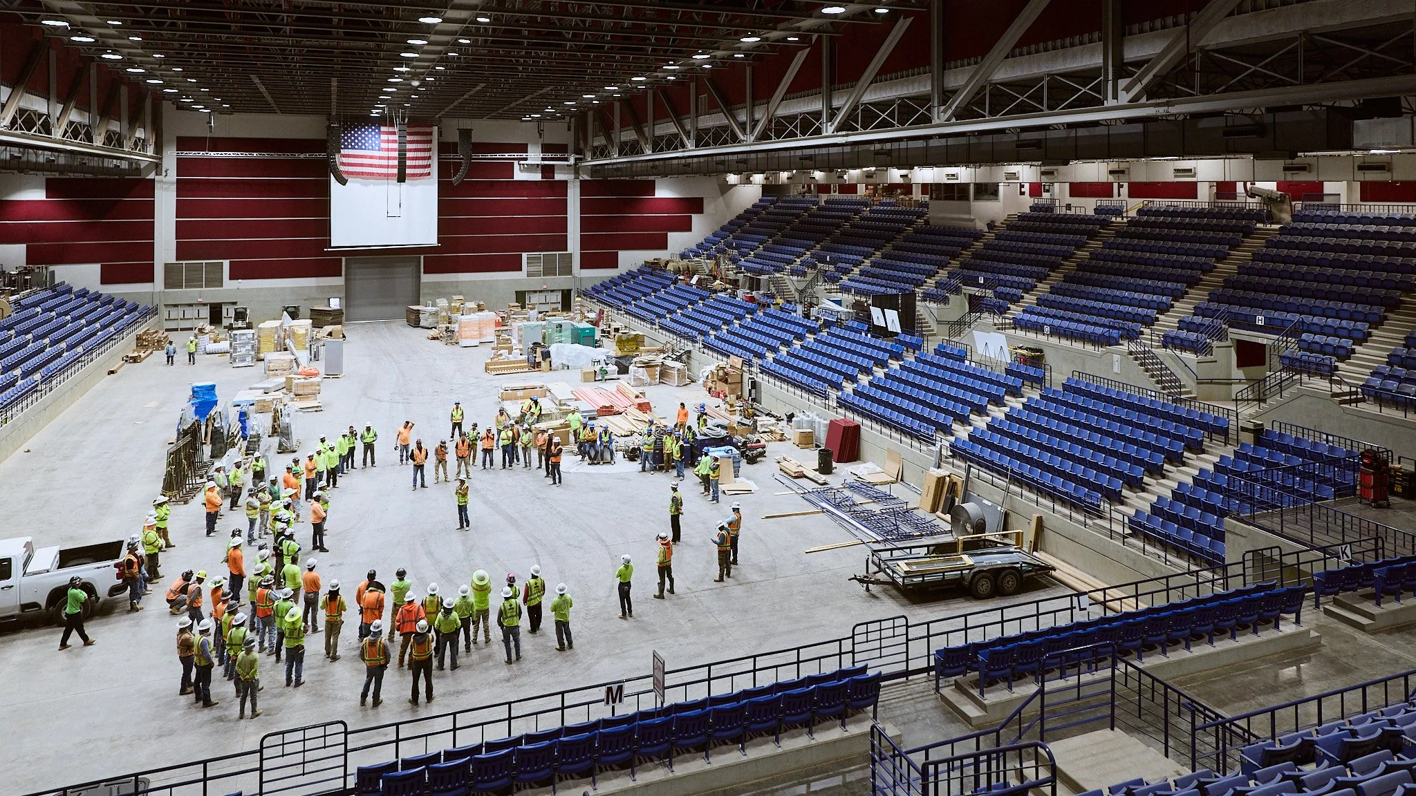 Construction workers and safety personnel working inside a large indoor arena with blue seating, a large American flag hanging above, and renovation or assembly materials scattered on the floor.