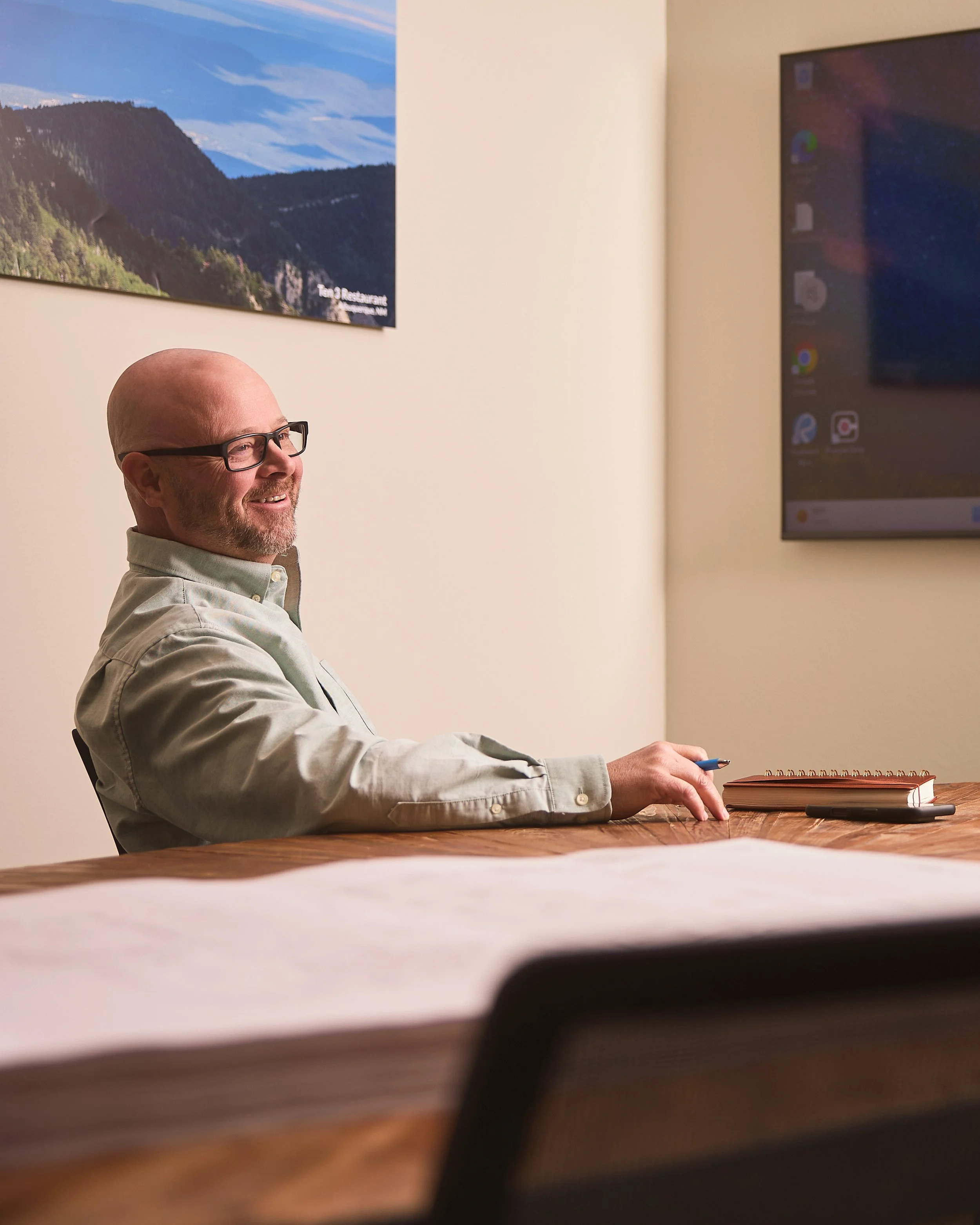 A man with glasses and a white shirt smiling and sitting at a wooden table with a closed notebook and pen in front of him in a conference or meeting room, with a world map or scenic poster on the wall and a large monitor to his right.