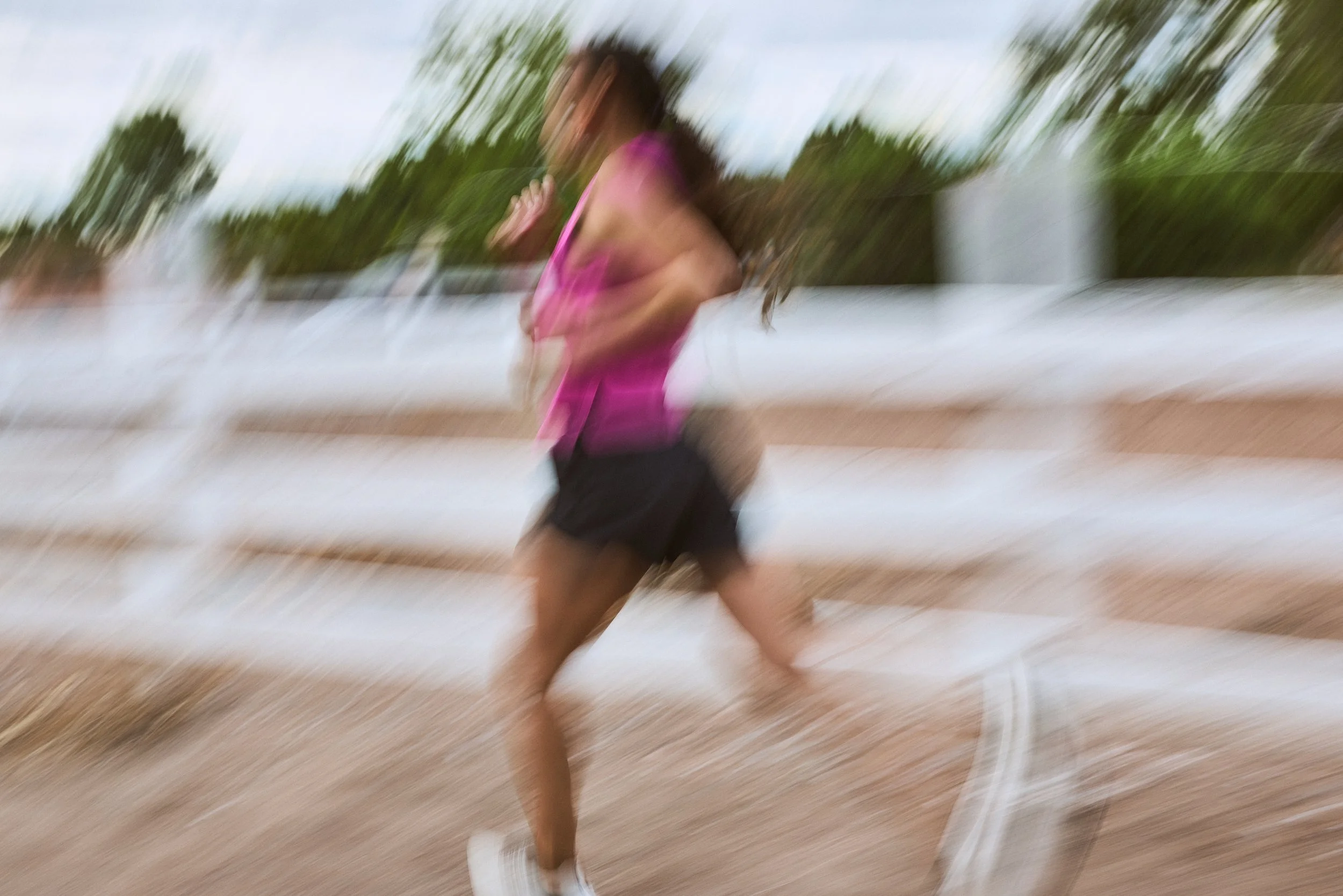 A woman running outdoors on a dirt path, wearing a pink tank top, black shorts, and white shoes, with trees and a white fence in the background.