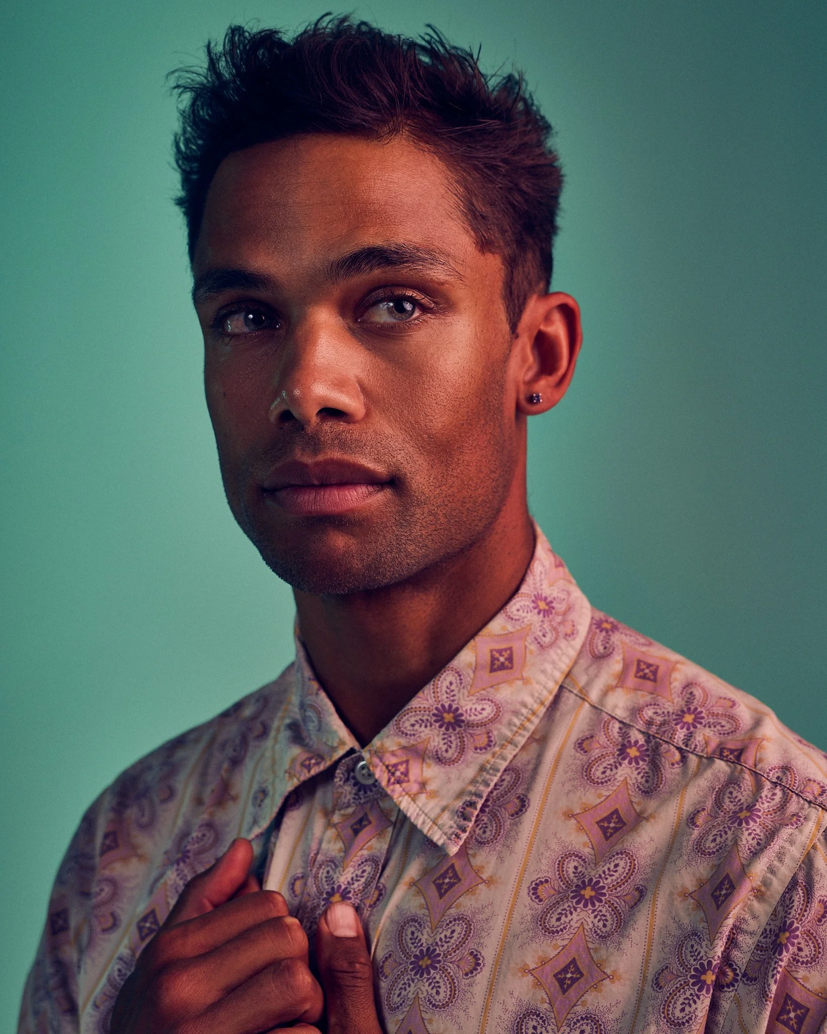 Close-up portrait of a young man with short, dark, and spiky hair, wearing a patterned shirt, holding his hand to his chest, set against a plain green background.