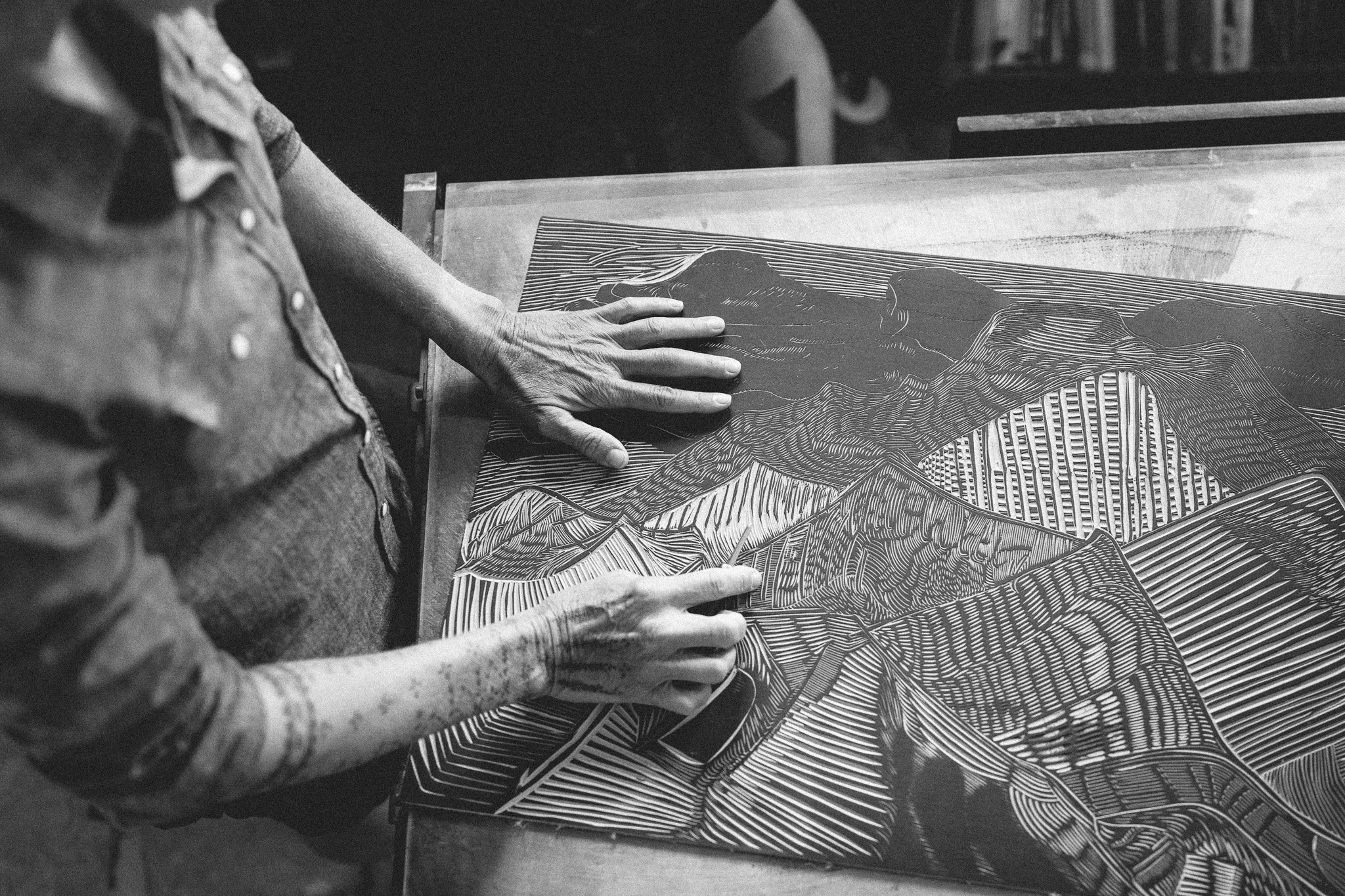 Black and white photo of an artist with a tattooed arm working on a detailed landscape print with mountains and lines.