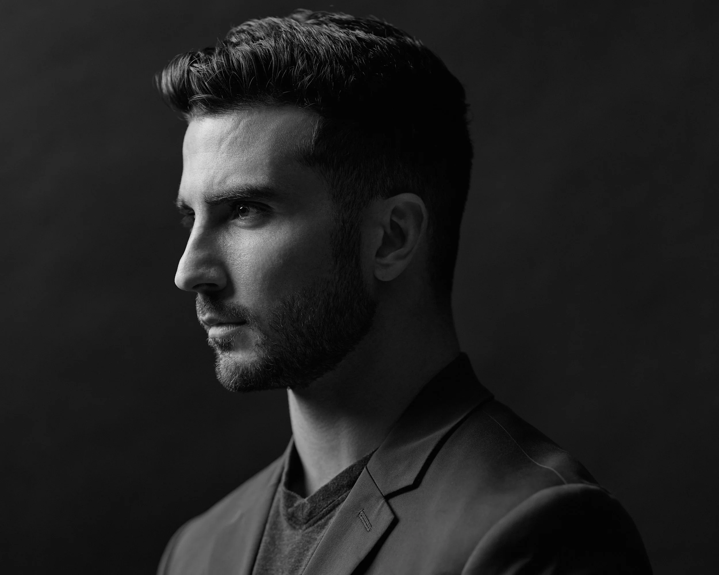 Black and white portrait of a young man with a styled haircut, wearing a jacket and a shirt, against a dark background.