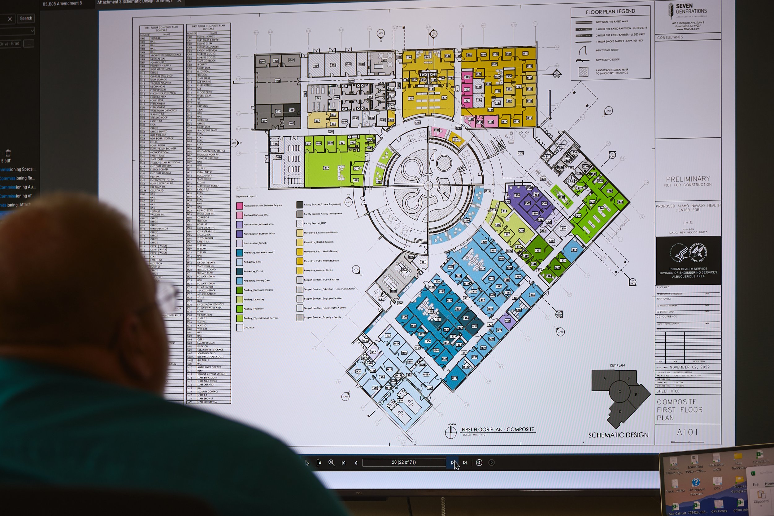 A detailed architectural floor plan displayed on a computer screen, showing various color-coded sections and labeled rooms for a proposed healthcare facility in Alamo, New Mexico, with a person observing it.