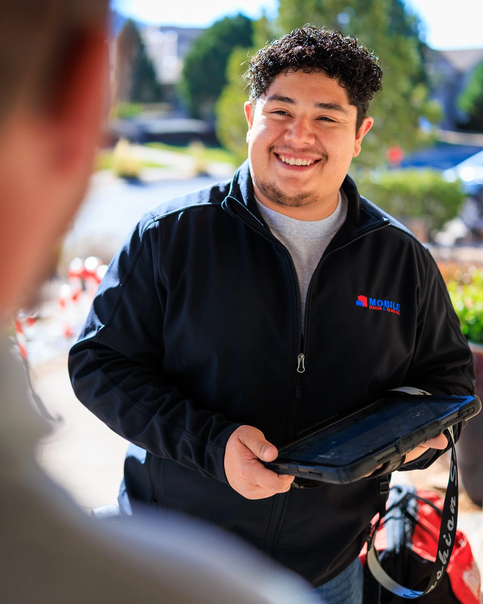 A young man with curly dark hair and a goatee smiling while holding a tablet outdoors. He is wearing a black jacket with a logo on the chest. There are trees and cars in the background, and the photo is taken on a sunny day.