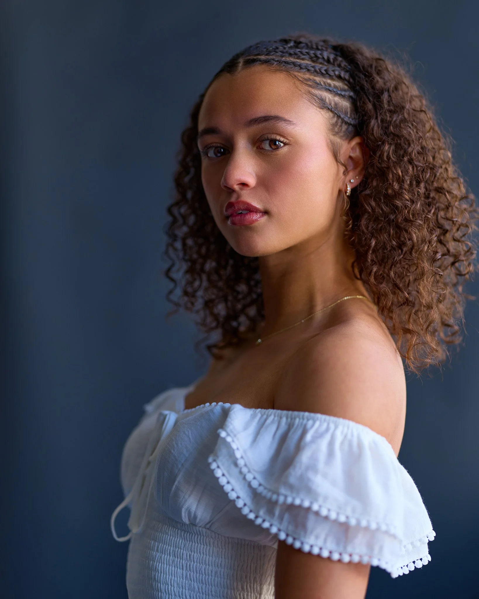 Close-up of a young woman with curly brown hair, wearing a white off-the-shoulder top with embroidered edges and earrings, looking at the camera against a dark blue background.