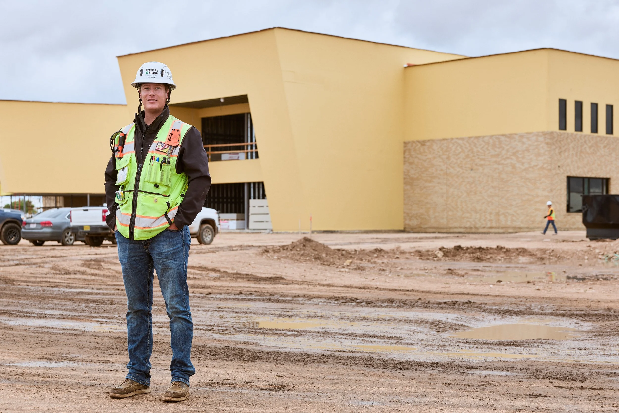 A construction worker wearing a white safety helmet and neon green reflective vest stands on a construction site with an unfinished building in the background. The site has dirt ground and parked vehicles. Another worker is visible in the distance.
