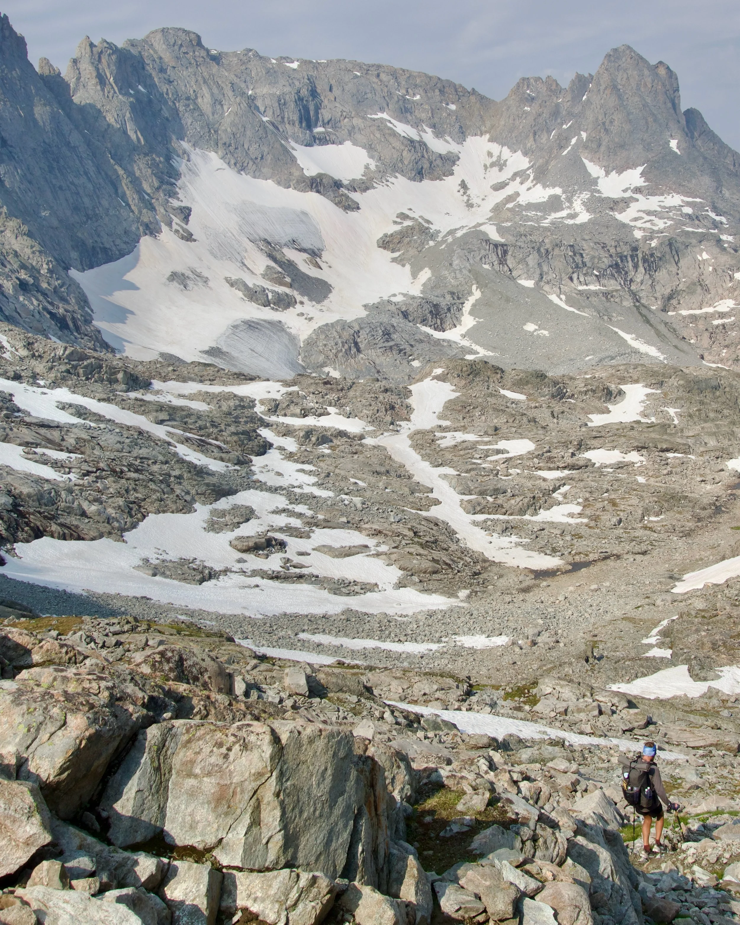 A rocky mountain landscape with patches of snow and a mountain climber in the bottom right corner carrying a backpack and walking among rocks.
