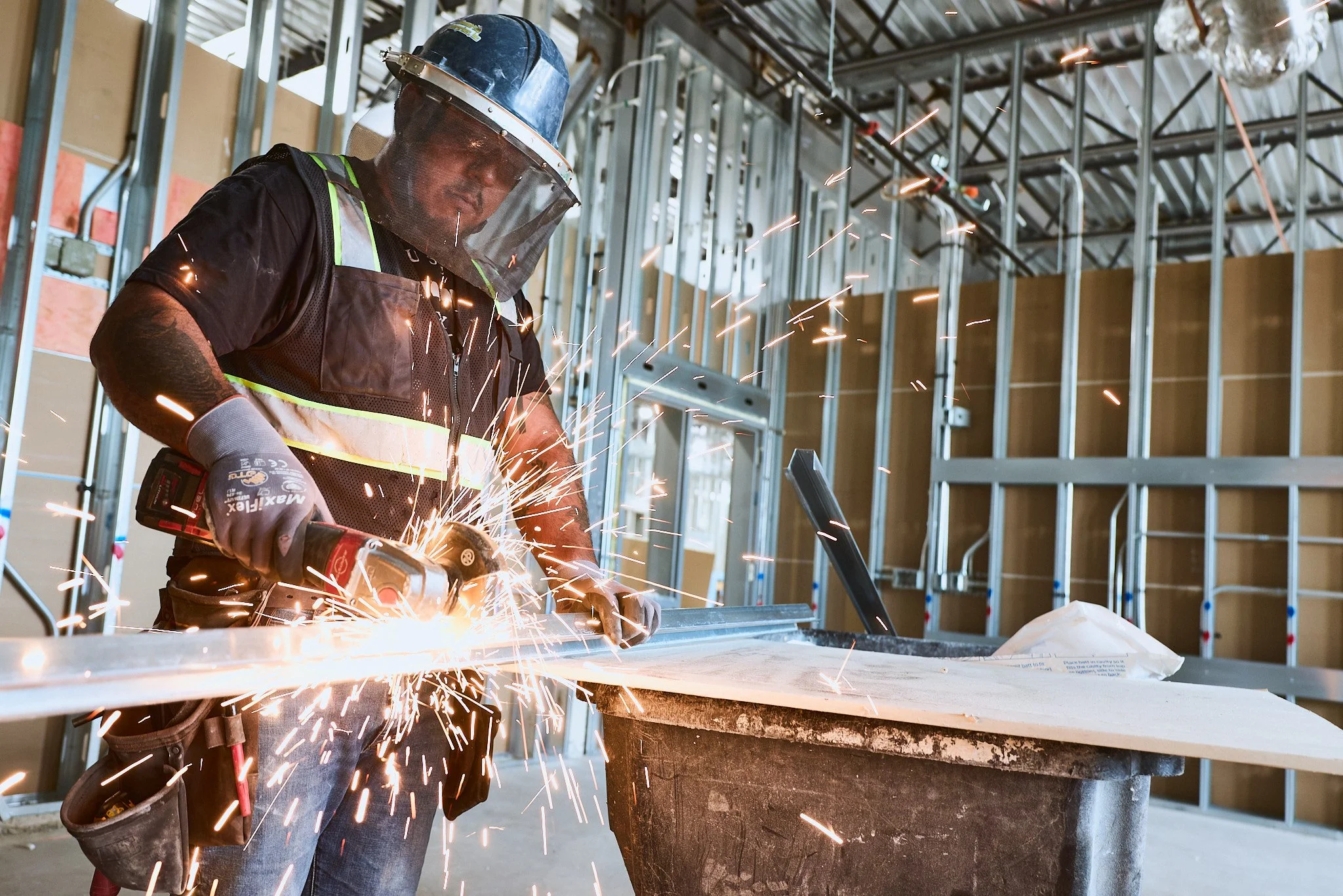 A construction worker wearing a helmet and protective gear is welding metal inside an unfinished building with exposed wall studs and a partially built countertop.