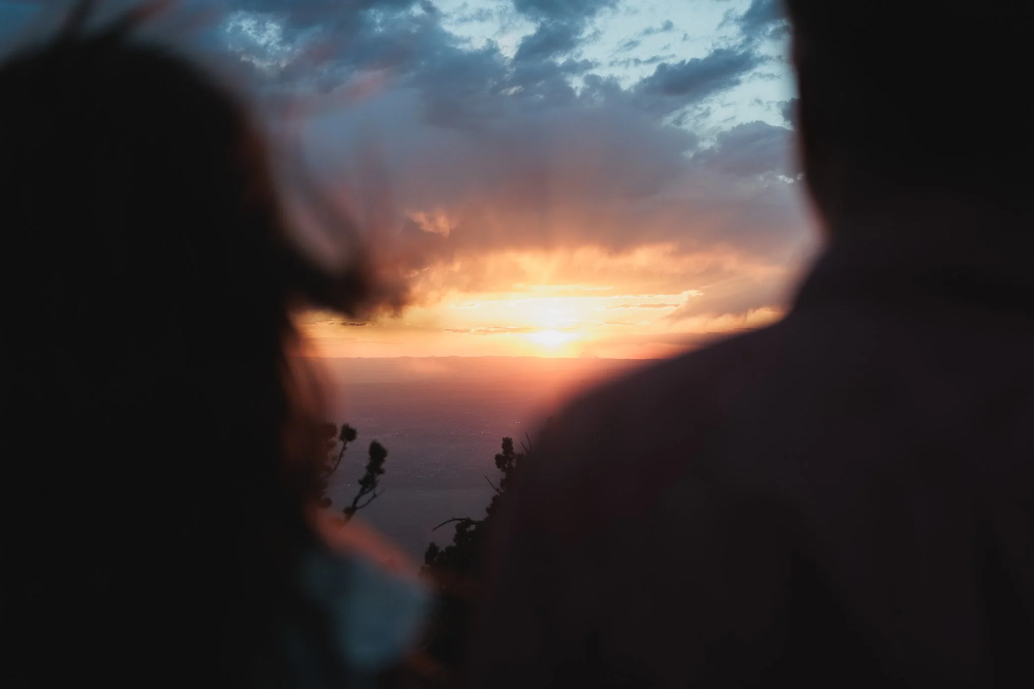 View of a sunset over the ocean, framed by dark silhouettes of trees and foreground objects, with clouds in the sky.