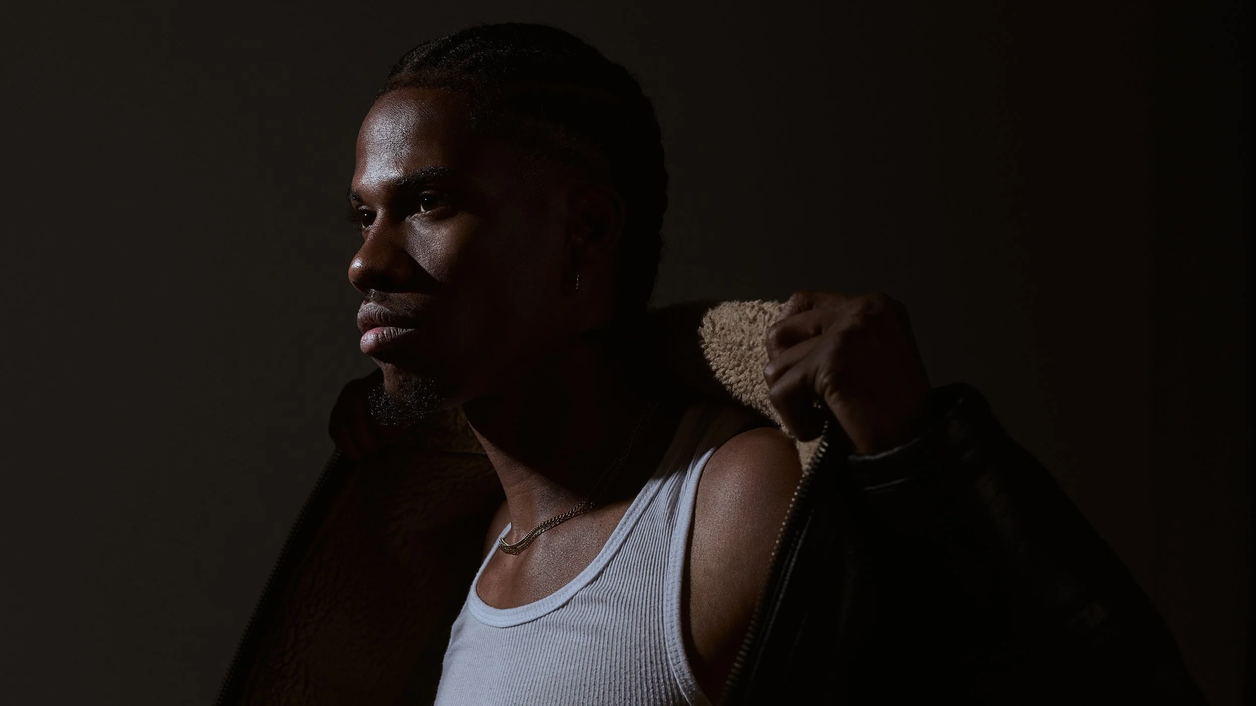 A man with dark skin and braided hair in a dark room, wearing a sleeveless shirt and a jacket with a furry collar.
