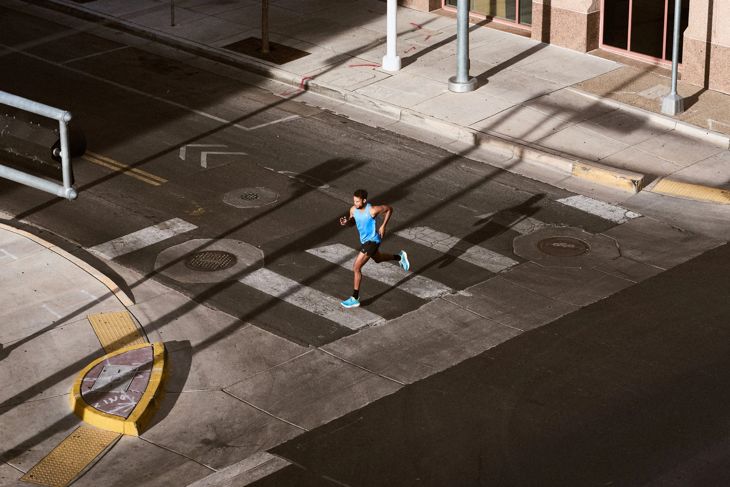 A man wearing a blue tank top and black shorts jogging on a city street at sunset with dark long shadows.