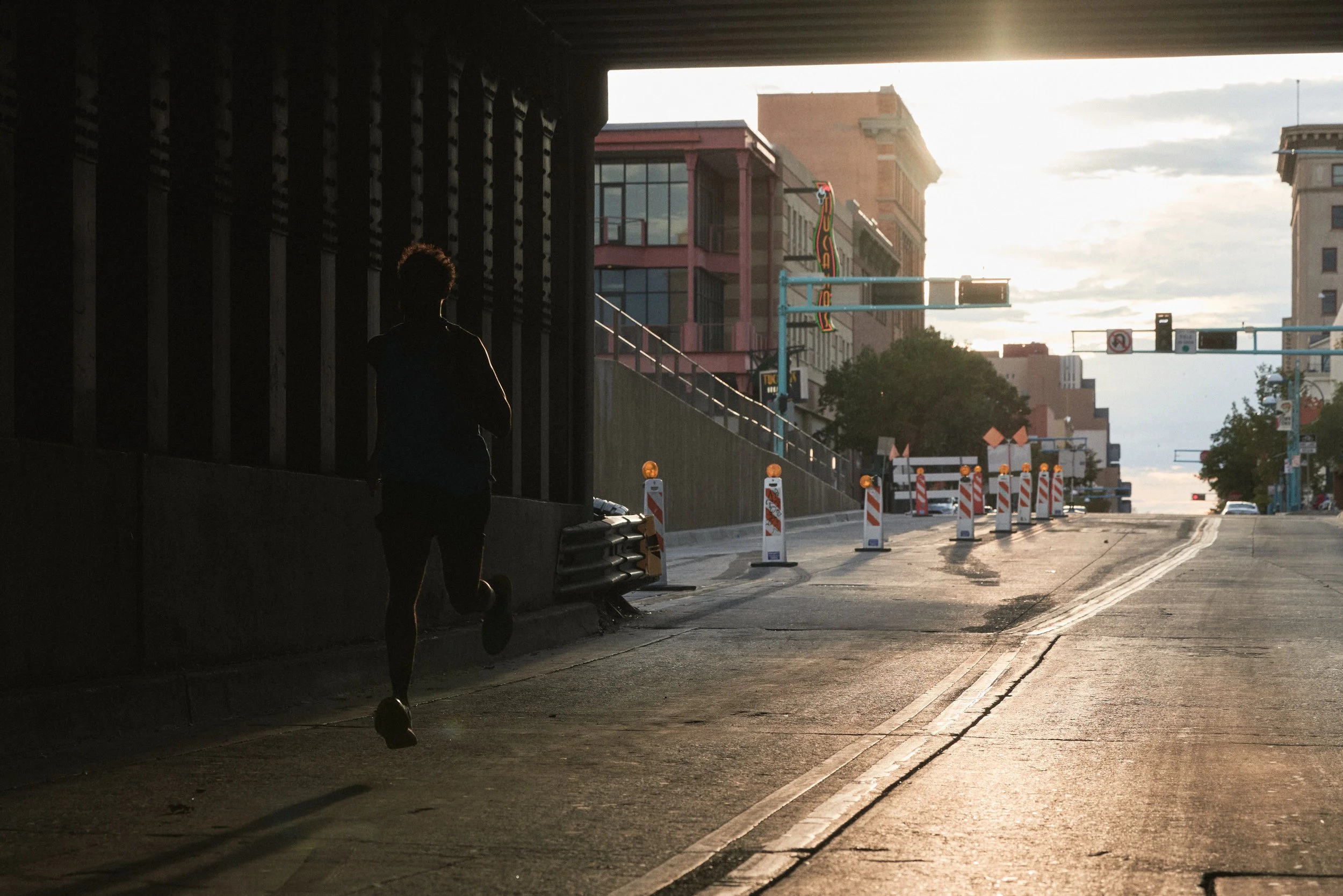 A person standing under an overpass on a city street during sunset, with construction barriers and buildings in the background.