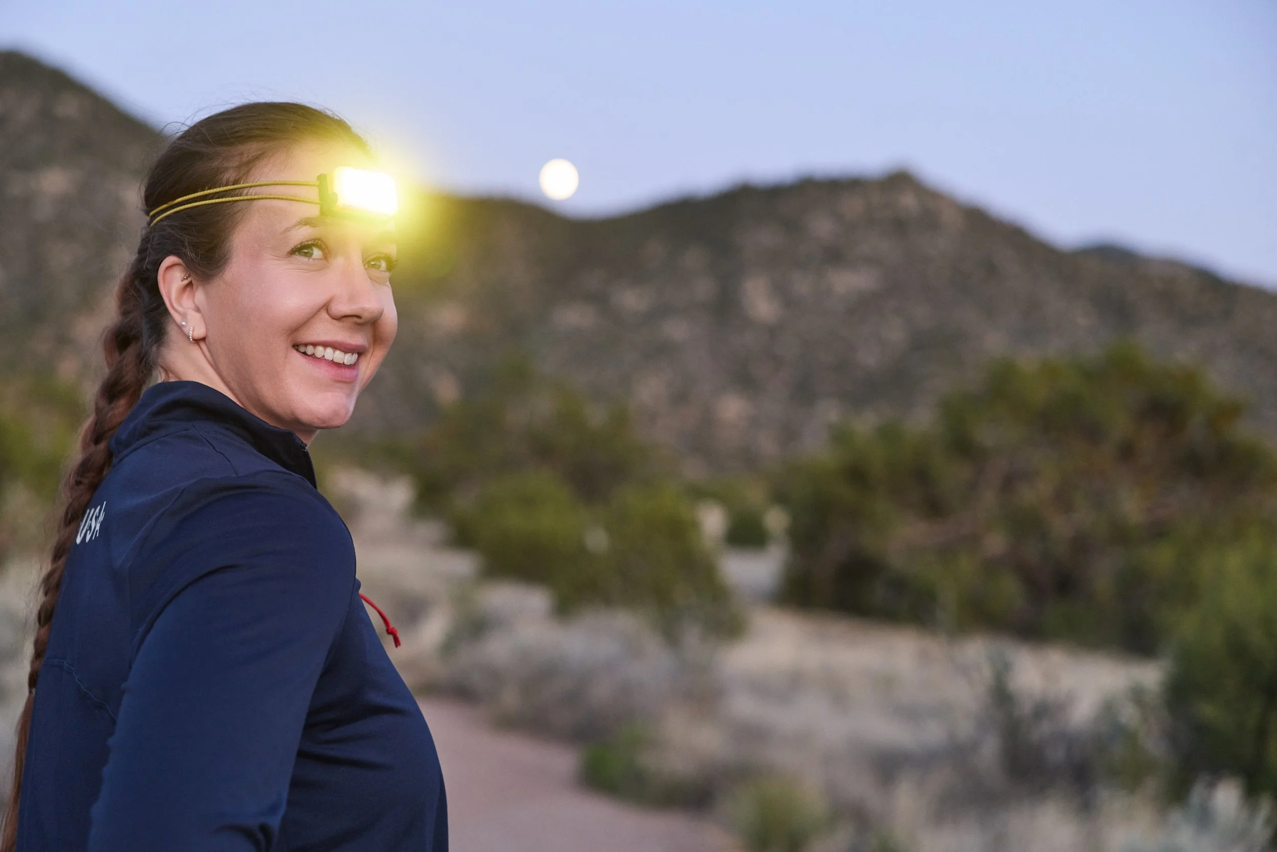 A smiling woman wearing a yellow headlamp and a navy blue jacket hiking outdoors at dusk with mountains and a creek in the background.