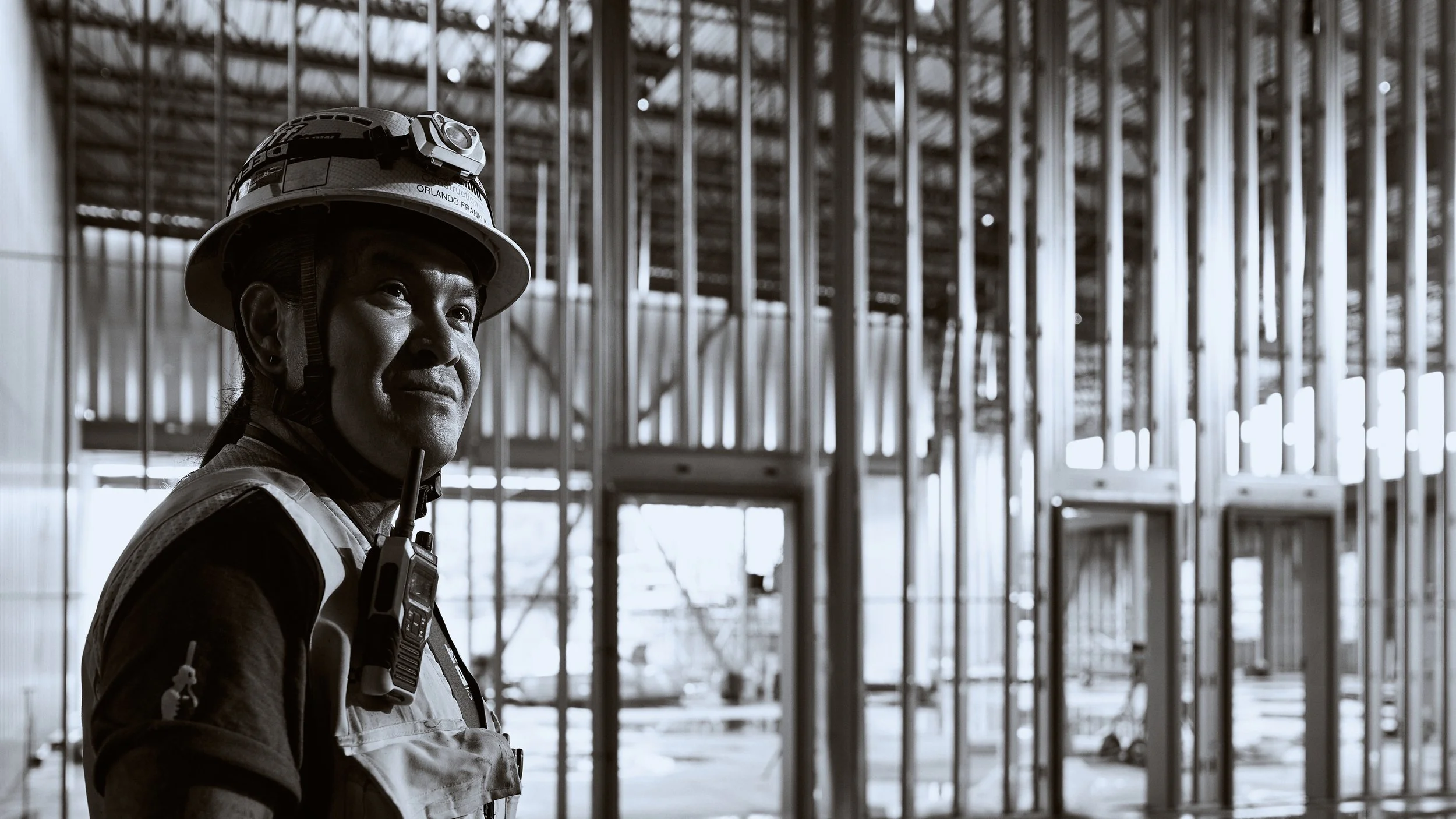 Construction worker wearing a helmet with a headlamp, standing inside a building under construction with exposed framing and steel beams.