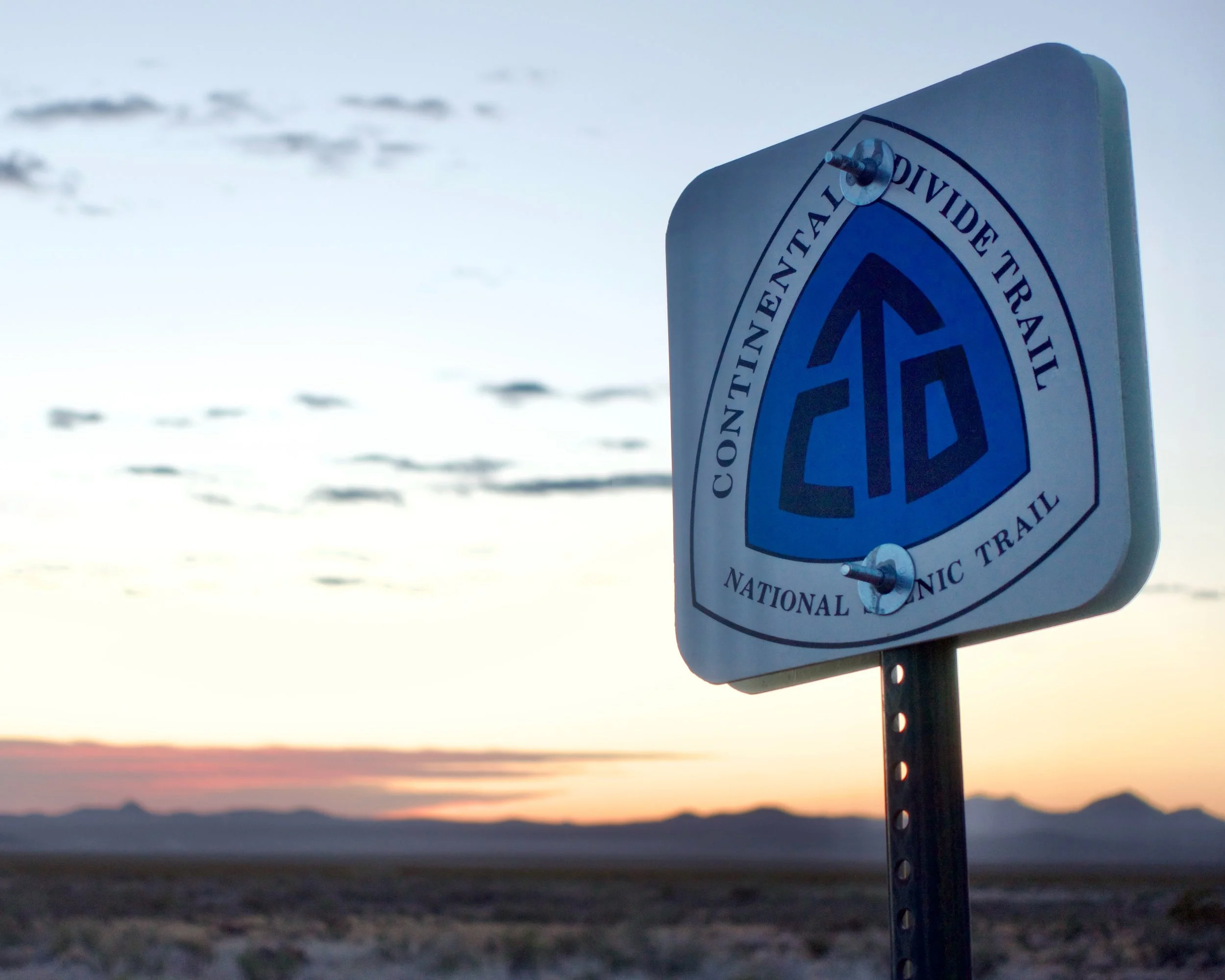 A sign for the Continental Divide Trail against a sunset sky with mountains in the distance.