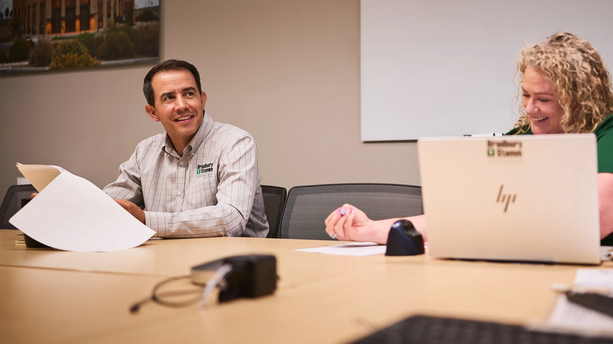Two people sitting at a conference table in a meeting room, smiling and talking. The man on the left is holding papers, while the woman on the right has a laptop in front of her. The background shows a framed picture and a whiteboard.