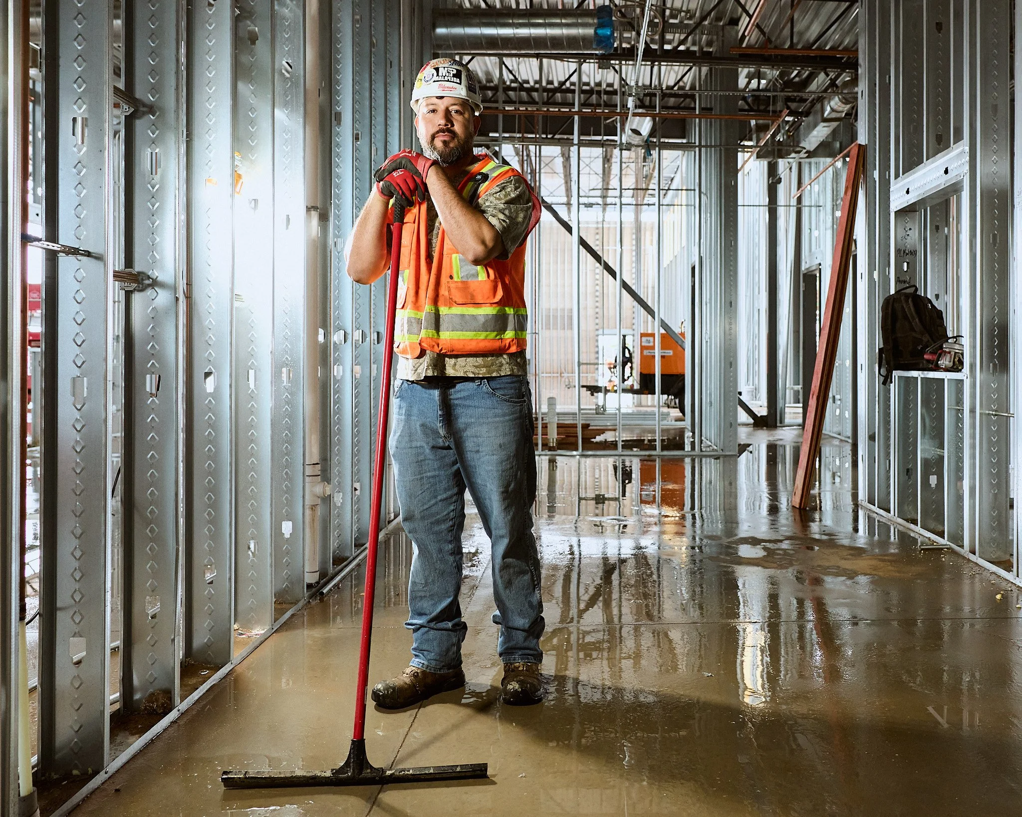 Man wearing construction safety helmet and orange safety vest standing with a shovel inside a partially built building framed with metal studs.