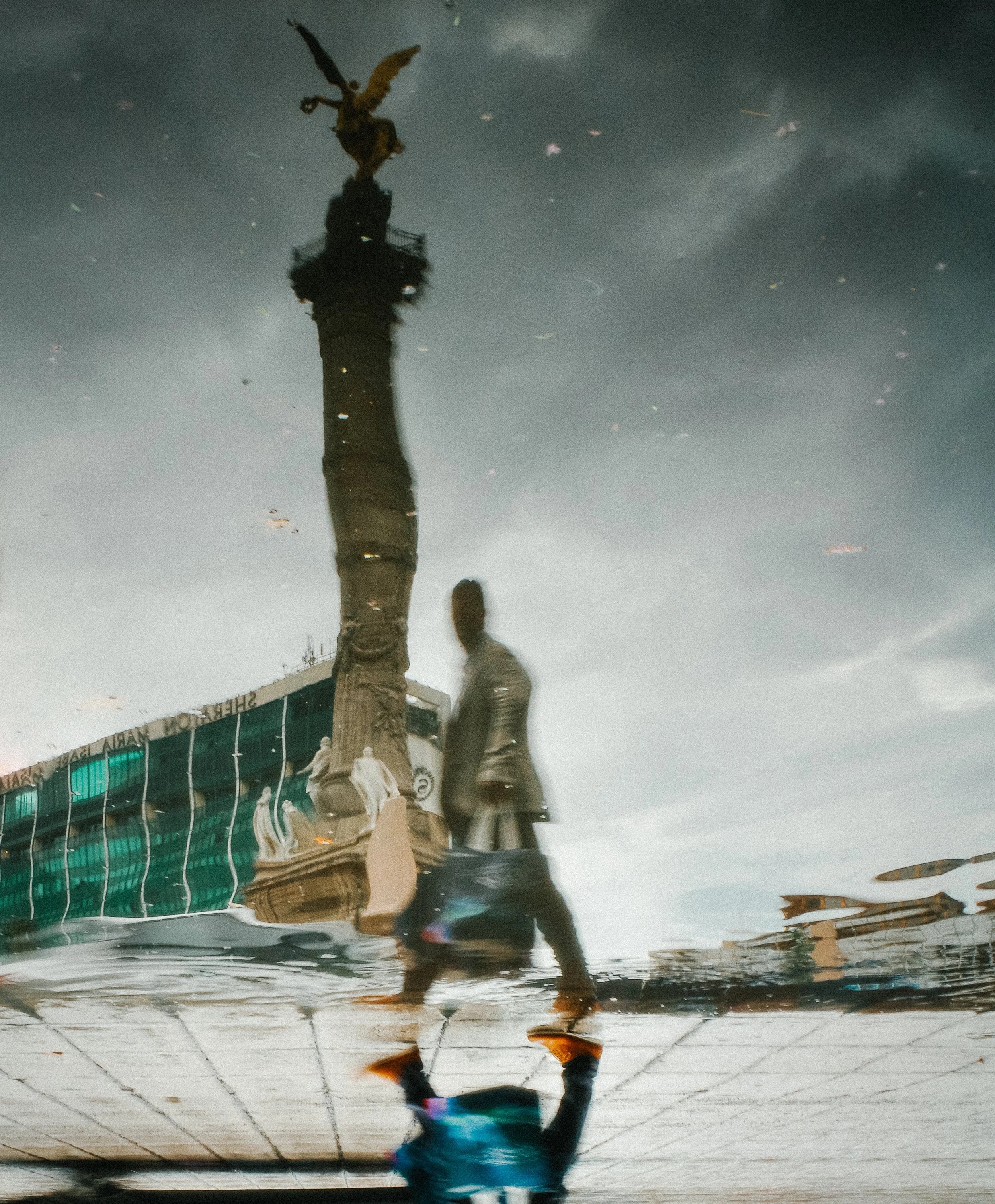 Reflection of a person walking on a wet cobblestone street with a building and a tree in the background, captured in a puddle, creating a mirror image.