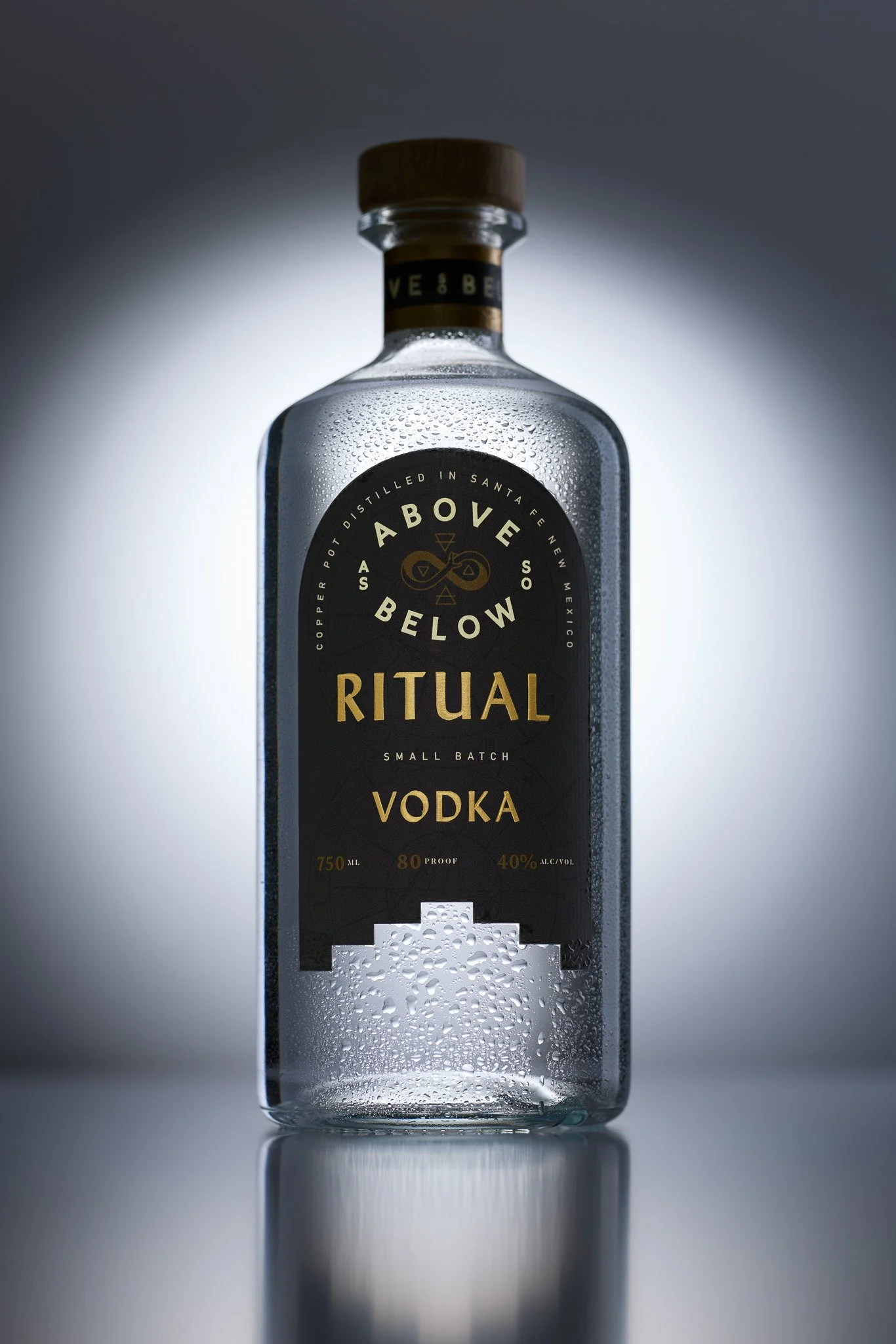 A clear glass bottle of Vodka with condensation and droplets on the surface, labeled as belonging to the 'As Above So Below' brand, with a black label and gold text, placed on a reflective surface with a gradient gray background.