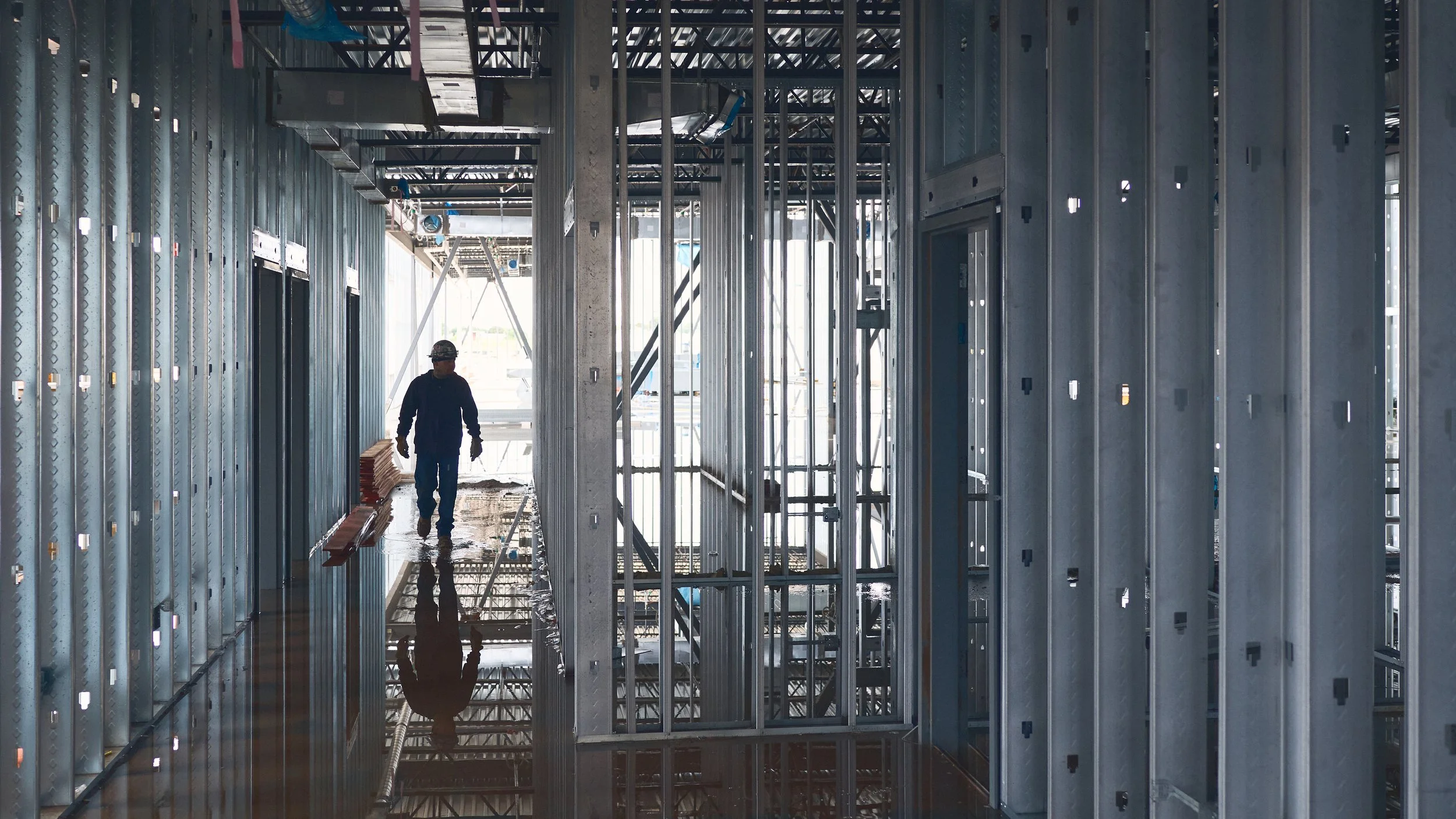 Construction worker wearing a helmet walking inside a building under construction with exposed metal framing and scaffolding.