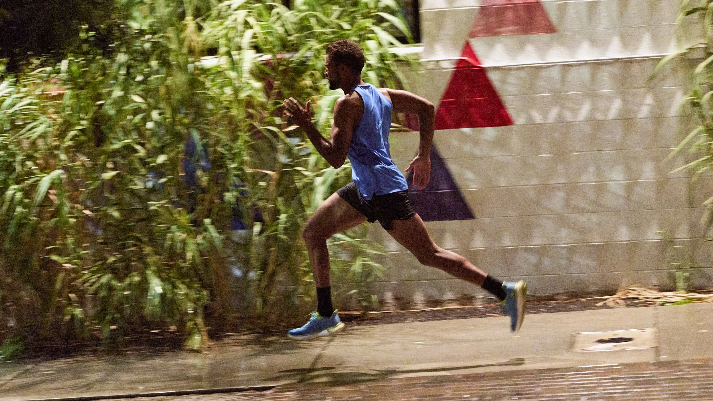 A man with dark skin running outside at night, wearing a blue sports tank top, black shorts, and blue running shoes, next to a concrete wall with a red and white striped pattern.