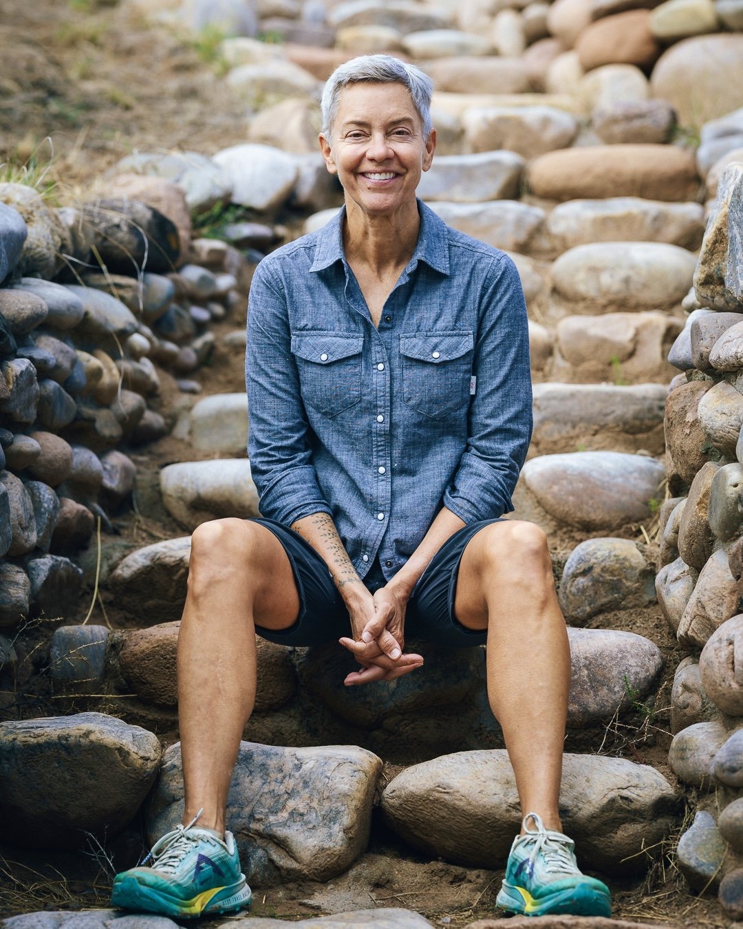 An older woman with short gray hair smiling, sitting on rocks and stairs made of stones outdoors, wearing a denim shirt, black shorts, and colorful sneakers.
