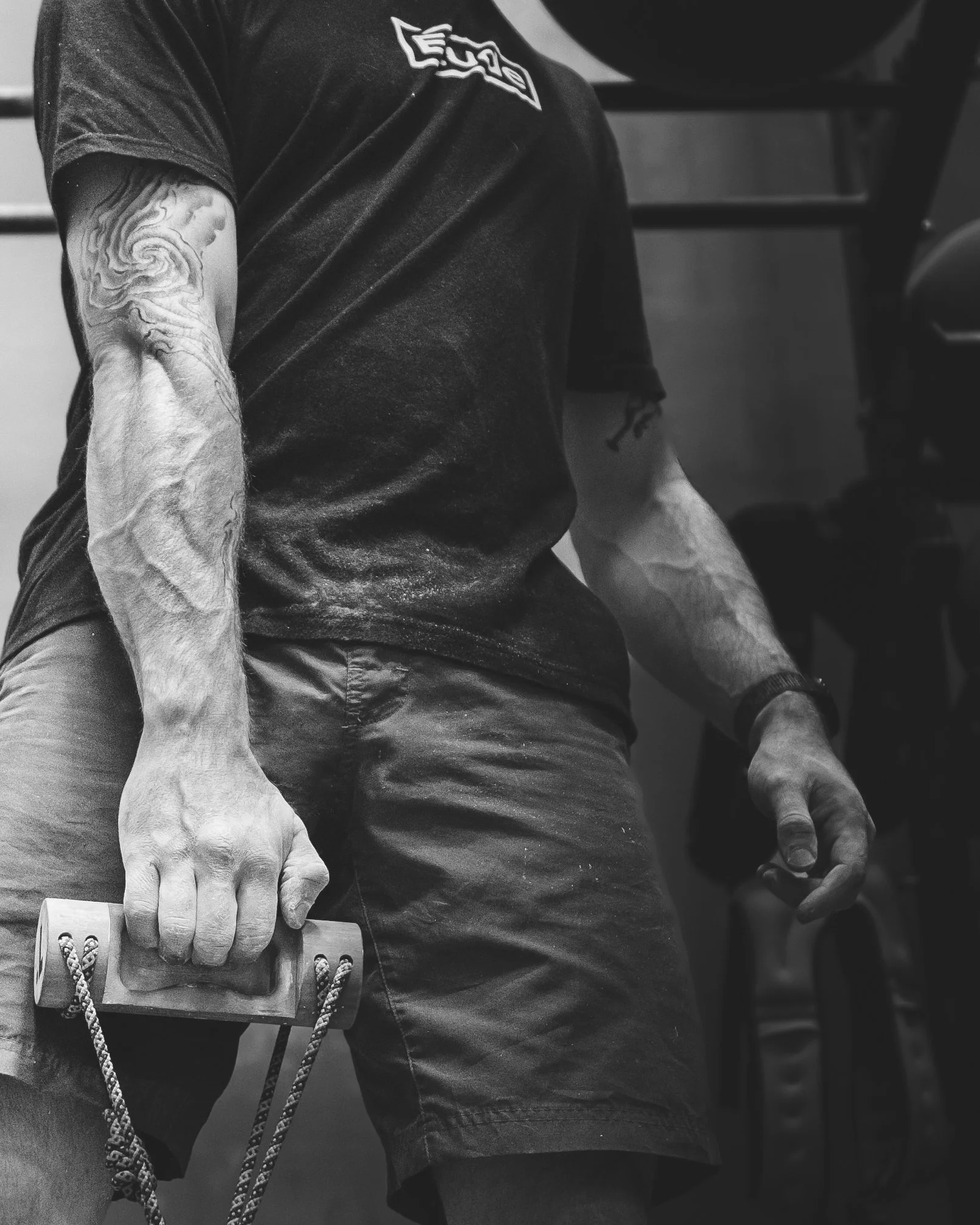 Close-up of a person's arm and hand gripping a wooden block with a rope attached, wearing a black T-shirt and dark shorts, in a gym or workout setting.