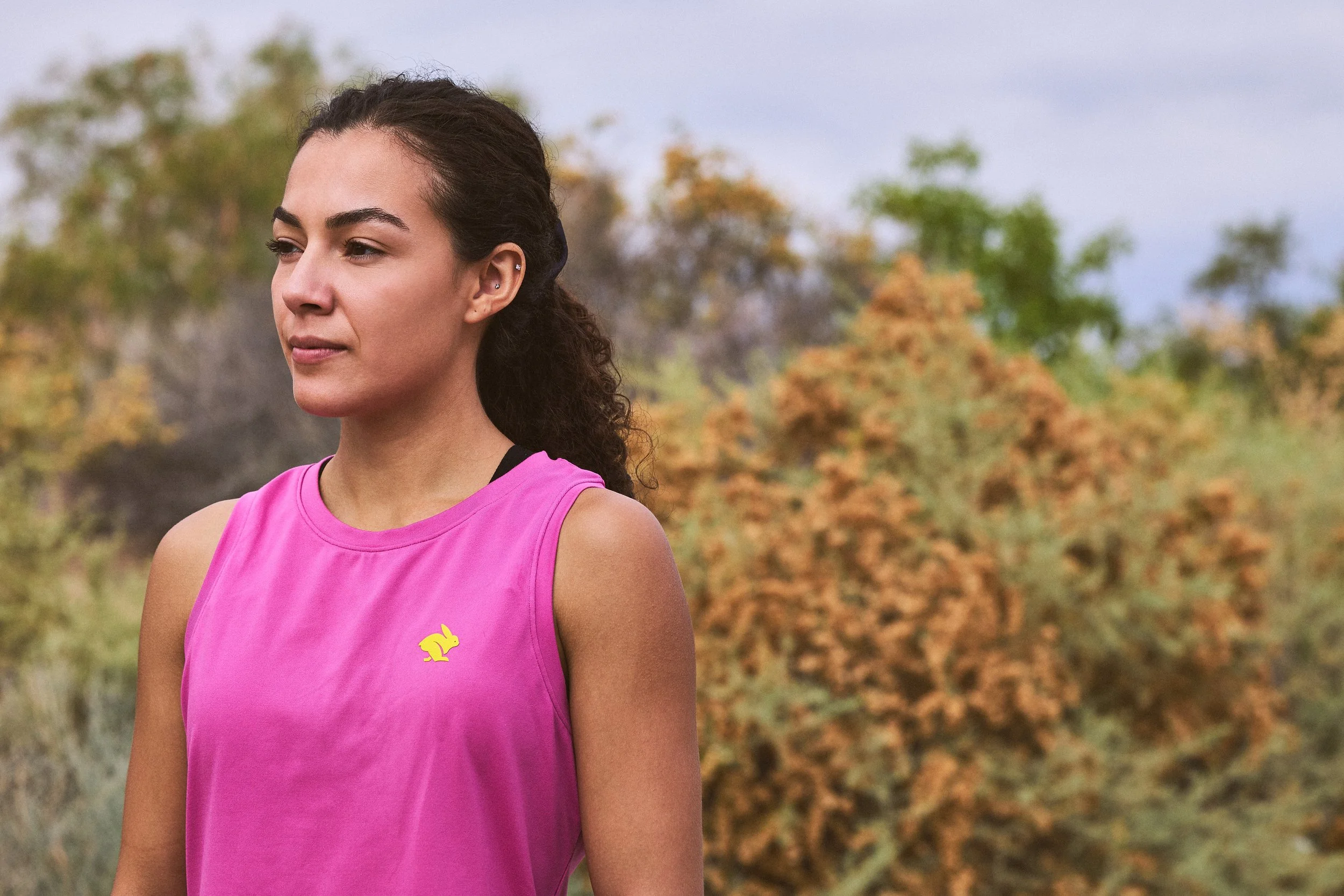 A woman in a pink athletic shirt outdoors, with trees and autumn foliage in the background.