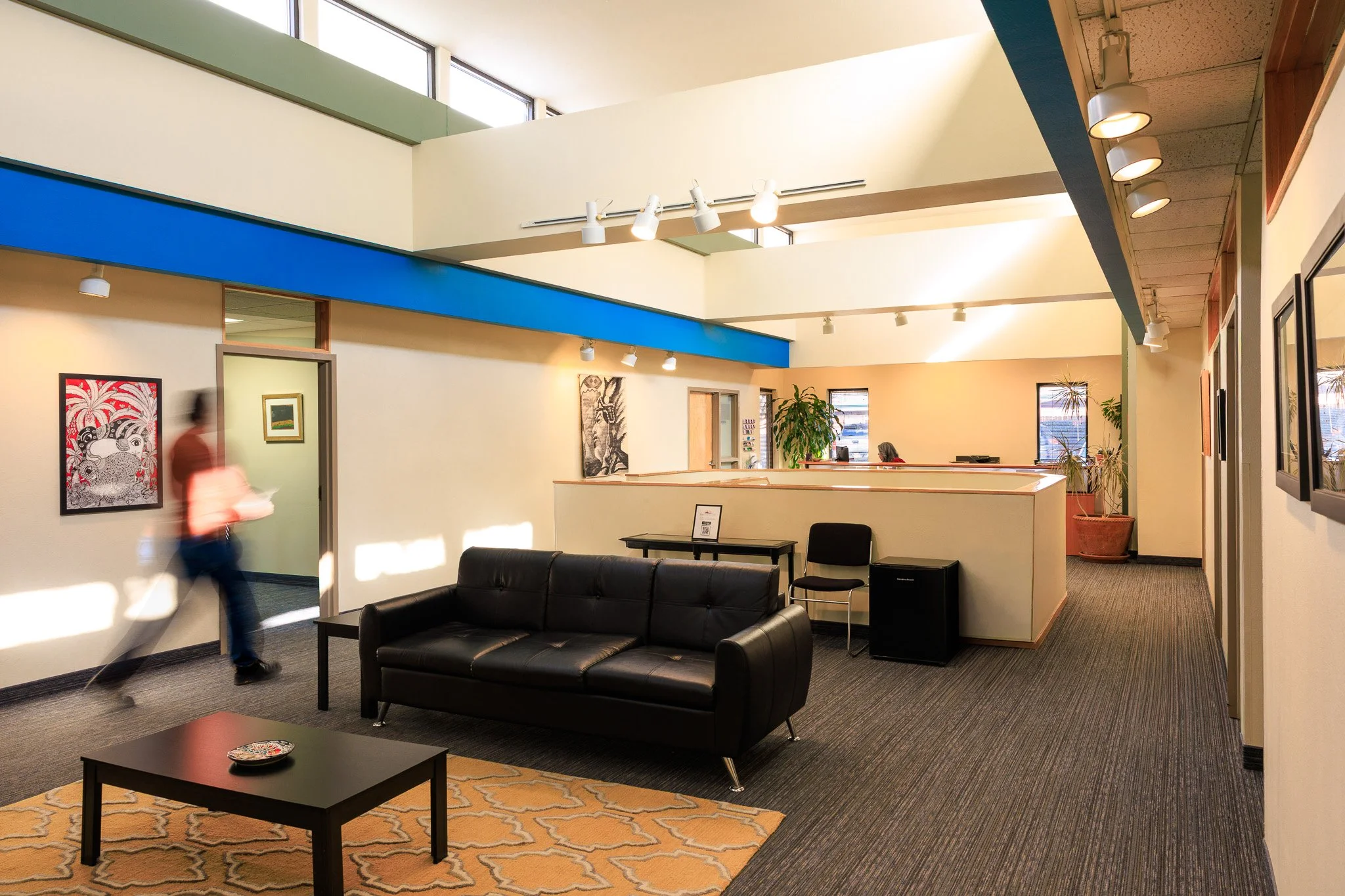 Lobby area with black leather sofa, coffee table, artwork on walls, and potted plants. Bright natural light from windows overhead.
