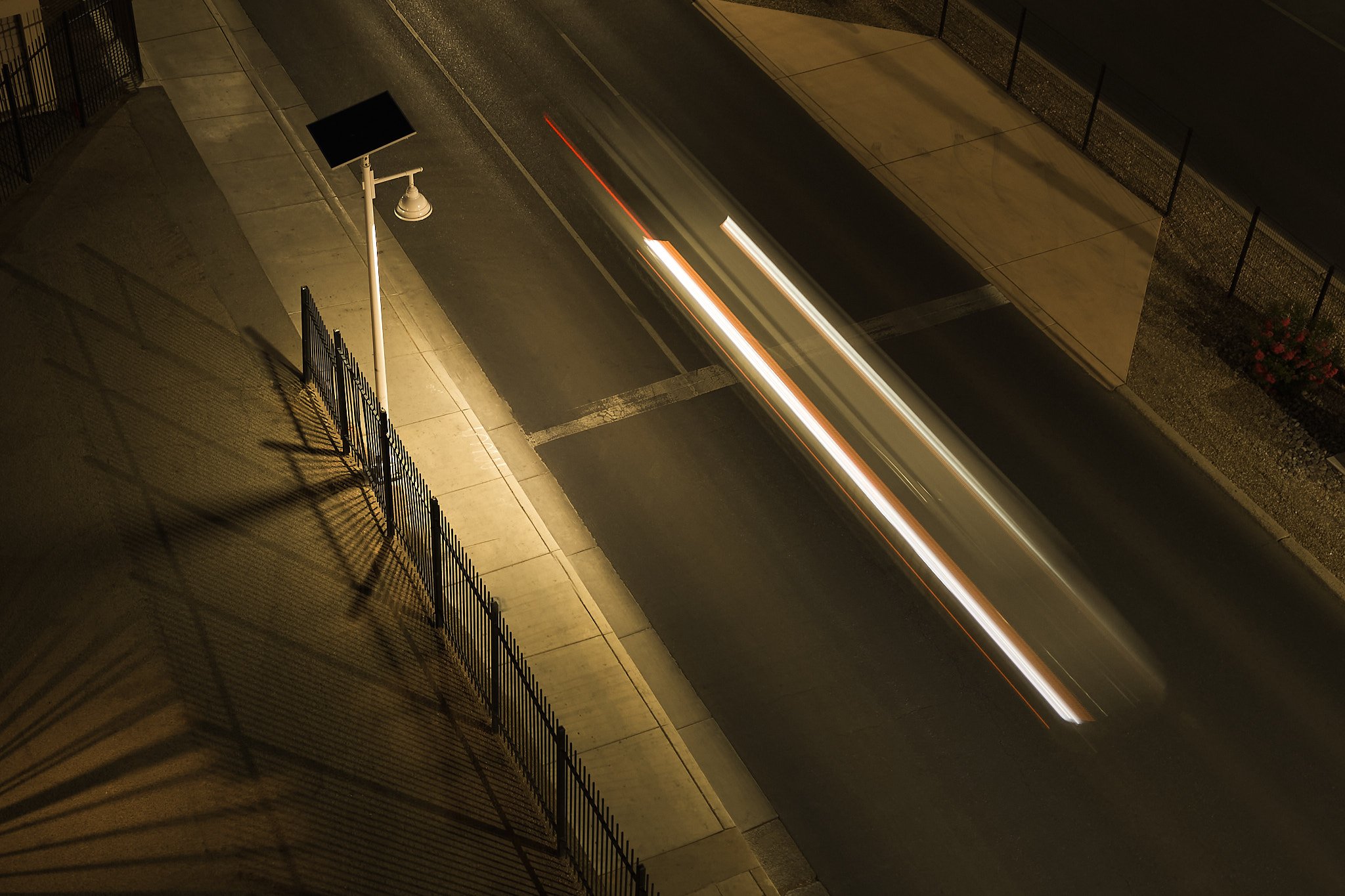 A nighttime street scene showing a sidewalk, a street fence, and a blurred vehicle passing by with light trails.