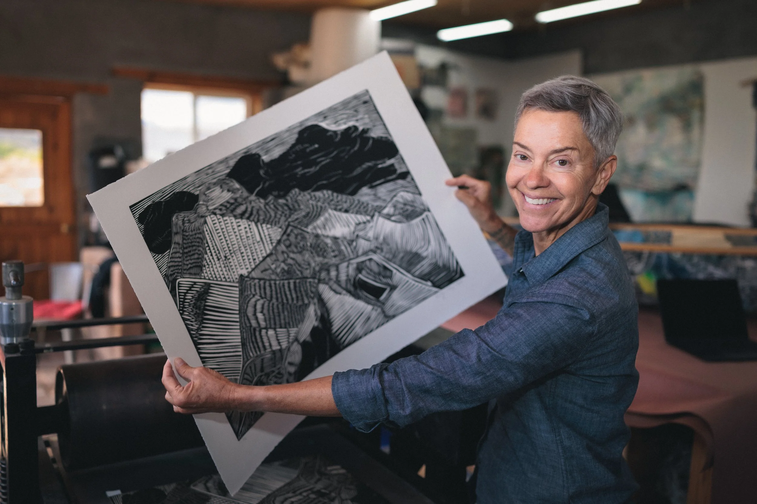A smiling woman with short gray hair holding a black and white artwork in an art studio.