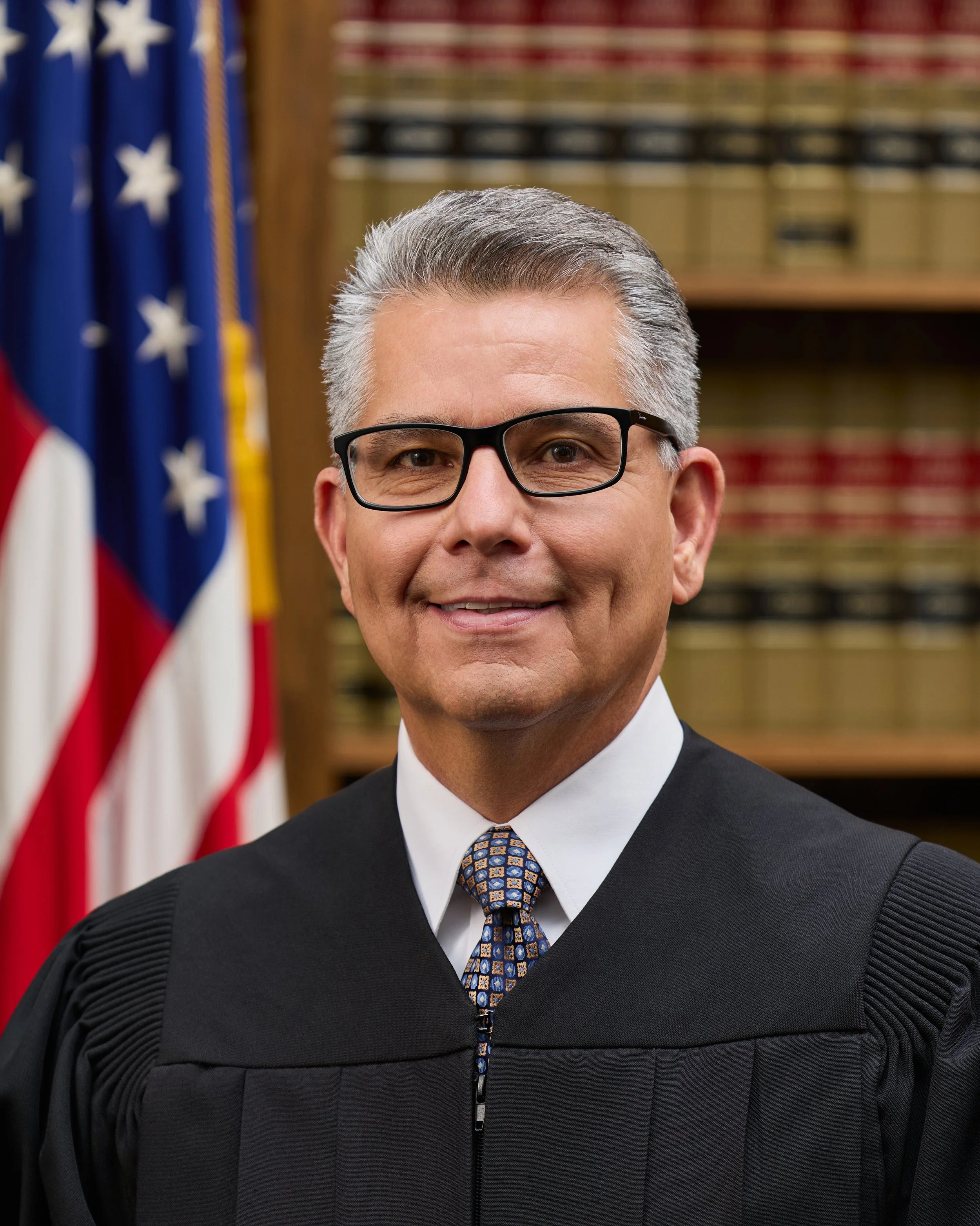 Close-up of a man with gray hair, glasses, wearing a black judicial robe, white shirt, and patterned tie, standing in front of American flags and a wooden bookshelf.