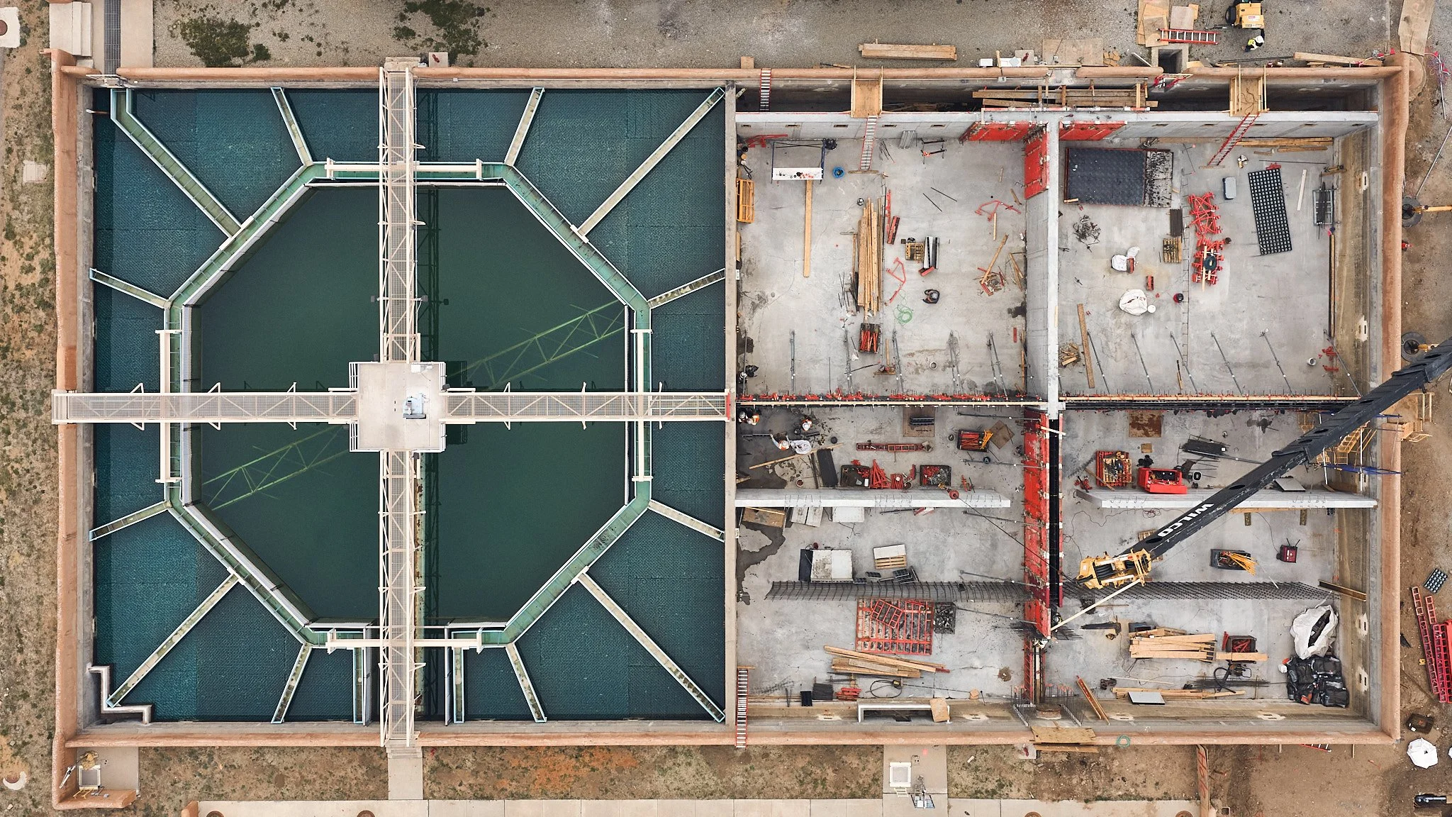 An aerial view of a construction site with a building under construction. The left side has a completed section with a green roof, the right side shows ongoing construction with building materials, machinery, and workers.