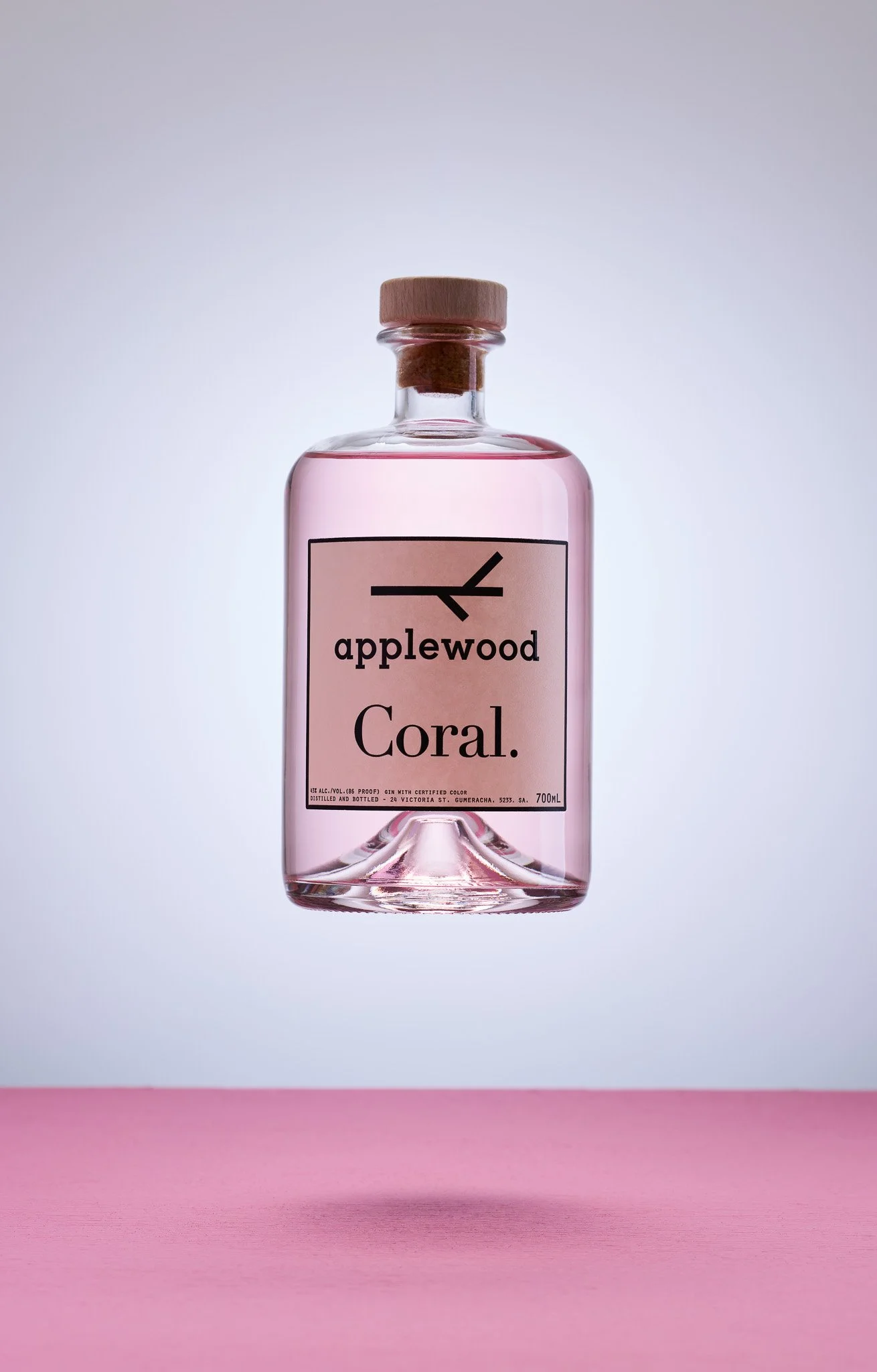 A pink-colored bottle of Applewood Coral gin with a wooden cap, floating above a pink surface against a light gray background.