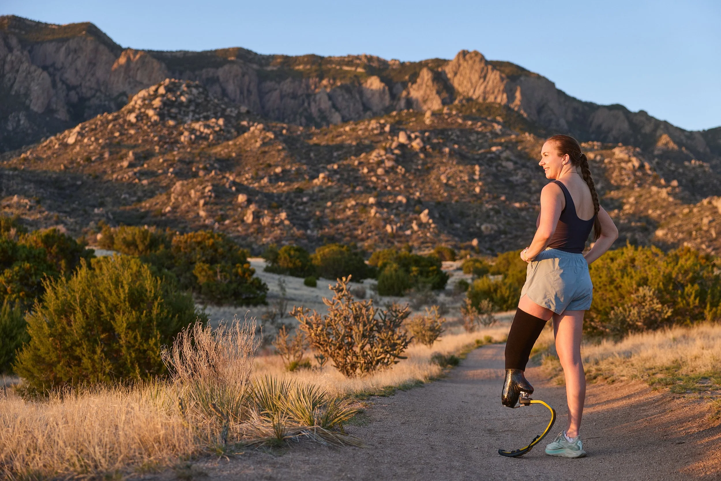 A woman standing on a trail in a mountainous desert landscape during sunset, wearing athletic clothing and roller skating.