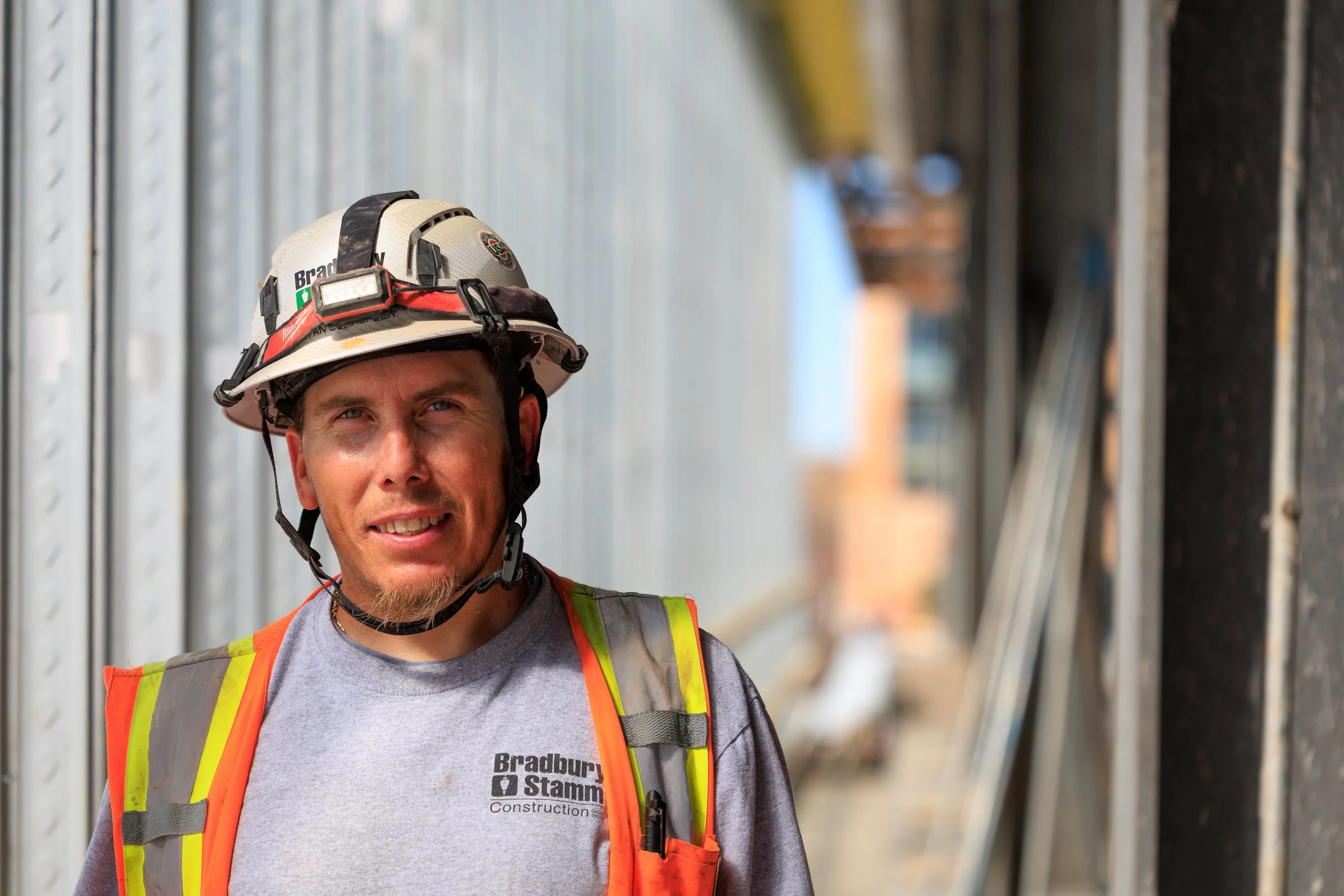 A construction worker wearing a grey shirt and orange safety vest, with a beige hard hat that has a headlamp and a name tag. The worker is smiling and standing next to a metal wall or structure.