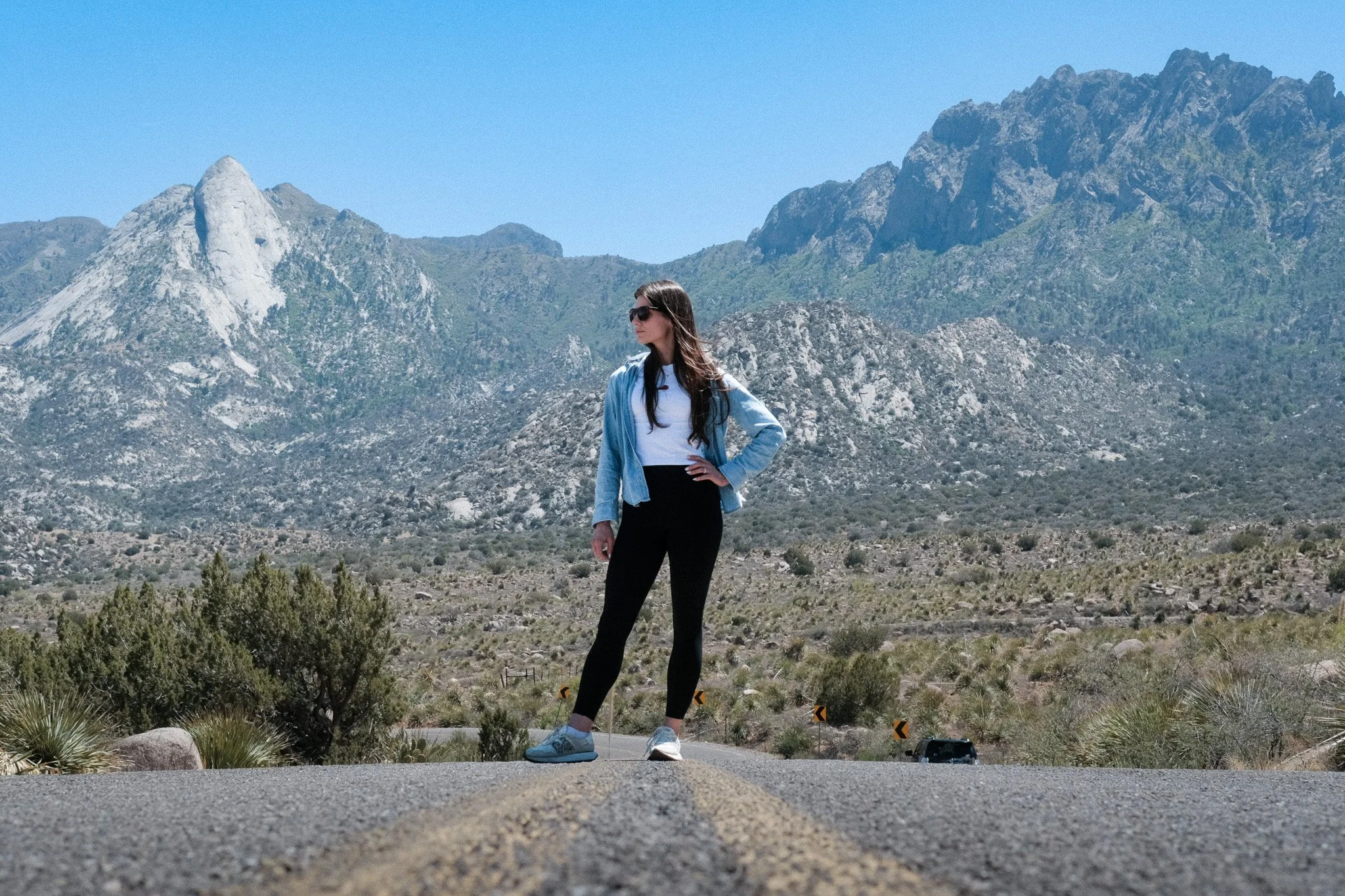 A woman wearing sunglasses, a white shirt, black pants, and sneakers standing on the side of a road in a mountainous desert area with mountains and a clear blue sky in the background.
