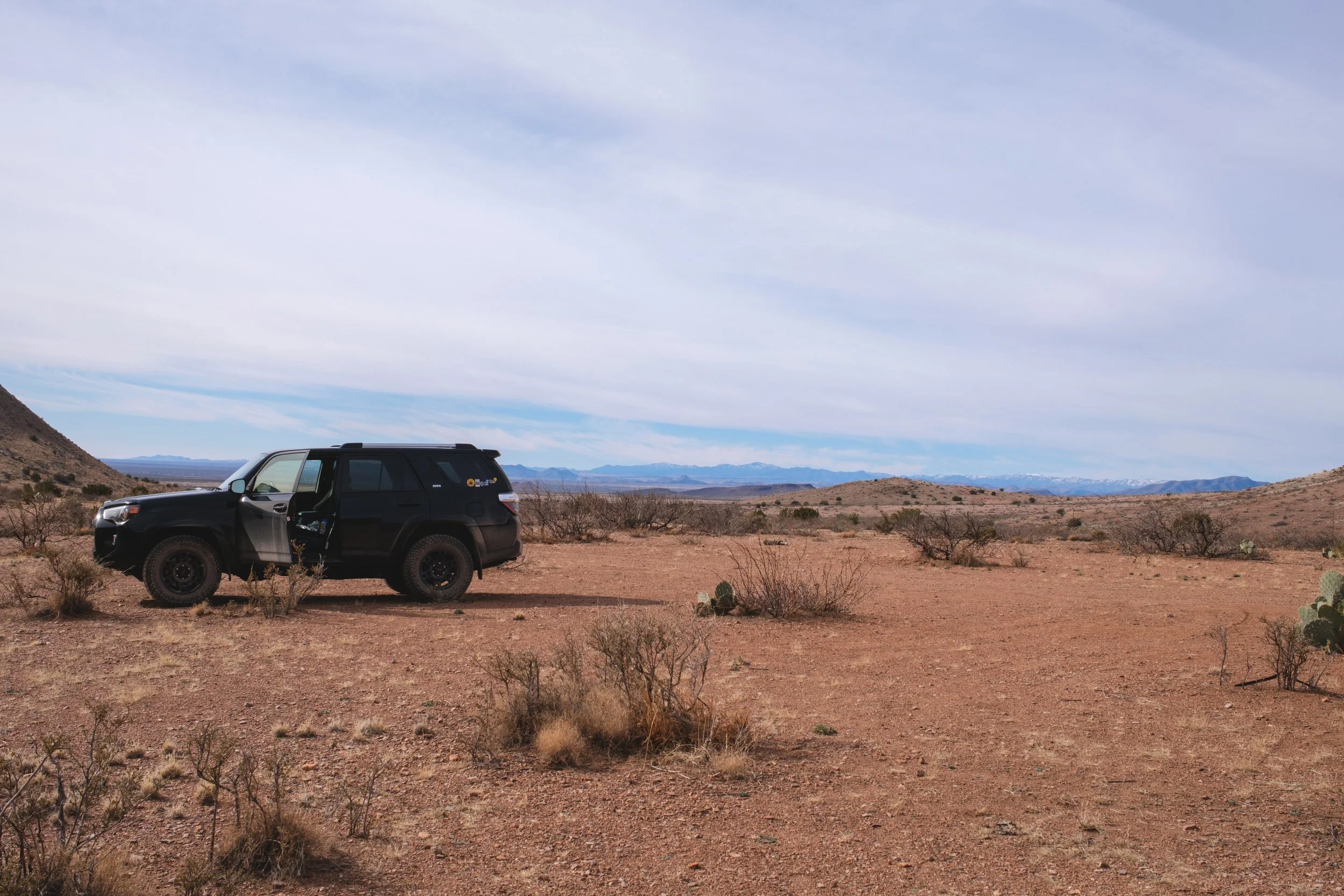 A black SUV parked on a desert landscape with mountains in the background and a cloudy sky overhead.