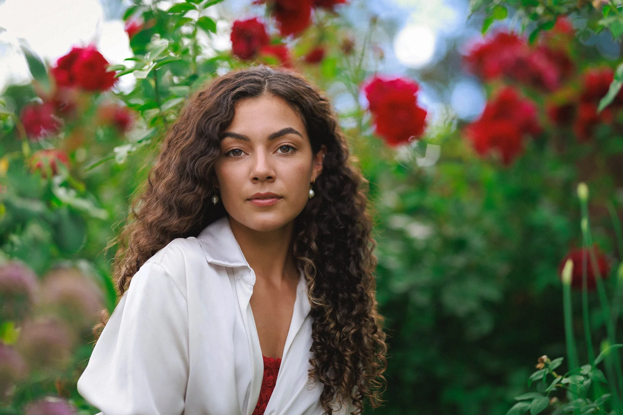 A woman with curly brown hair and pearl earrings standing among red flowers and green foliage.