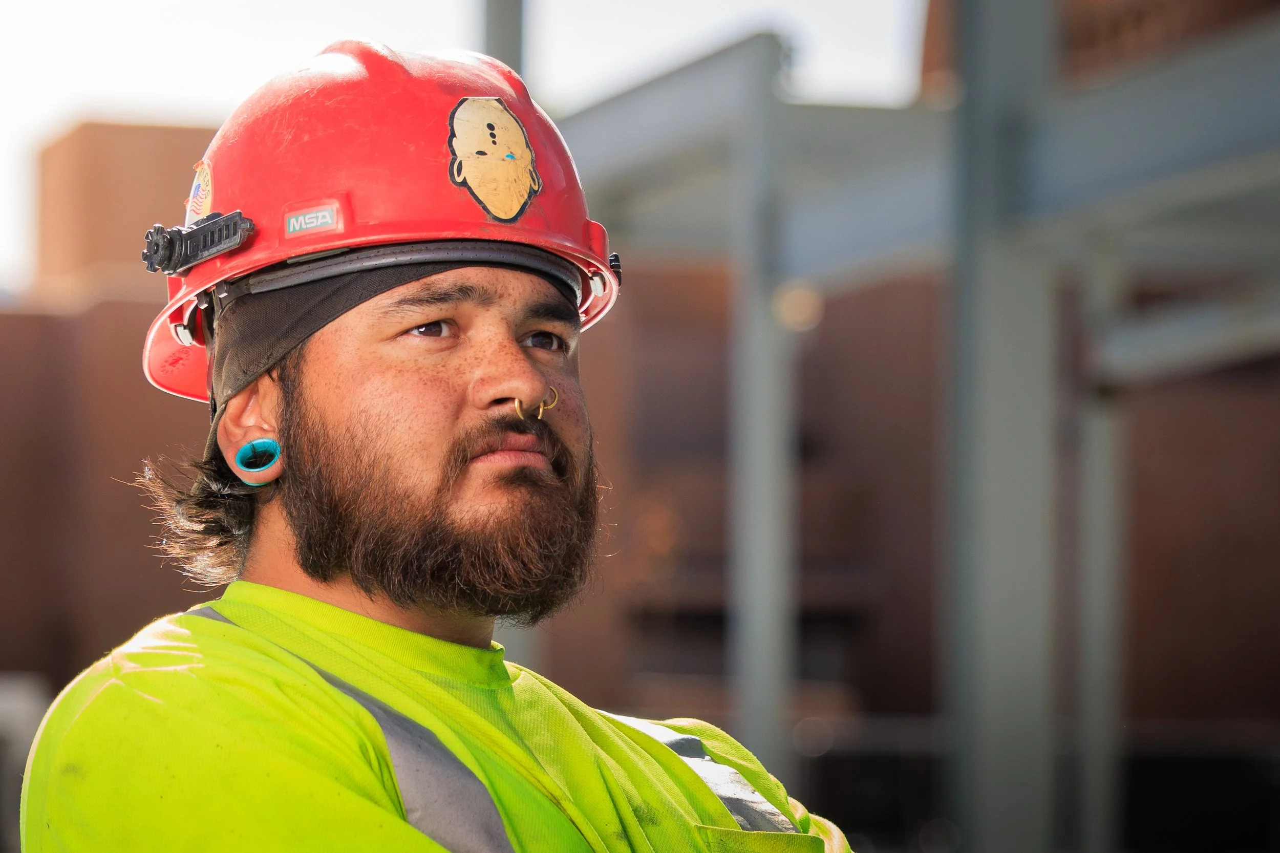 Close-up of a construction worker wearing a red safety helmet with a sticker, a black headband, a tiara piercing, a septum piercing, and teal earrings, dressed in a neon yellow safety shirt.