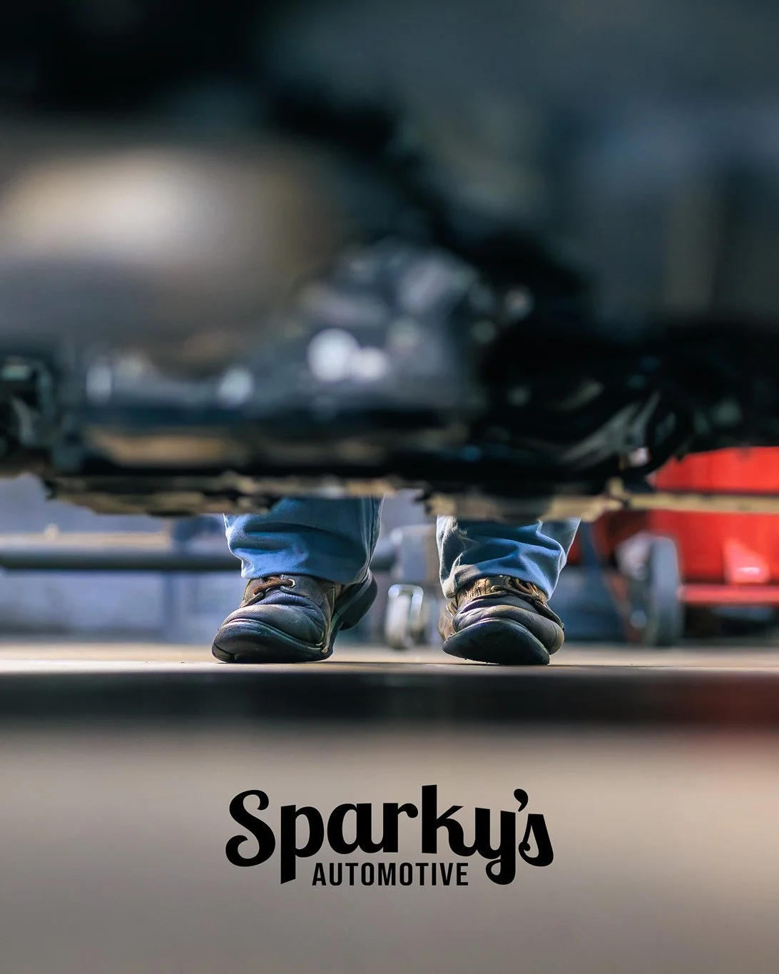 Customer's perspective of a car from underneath at Sparky's Automotive, showing the person's shoes and jeans with other cars in the background.