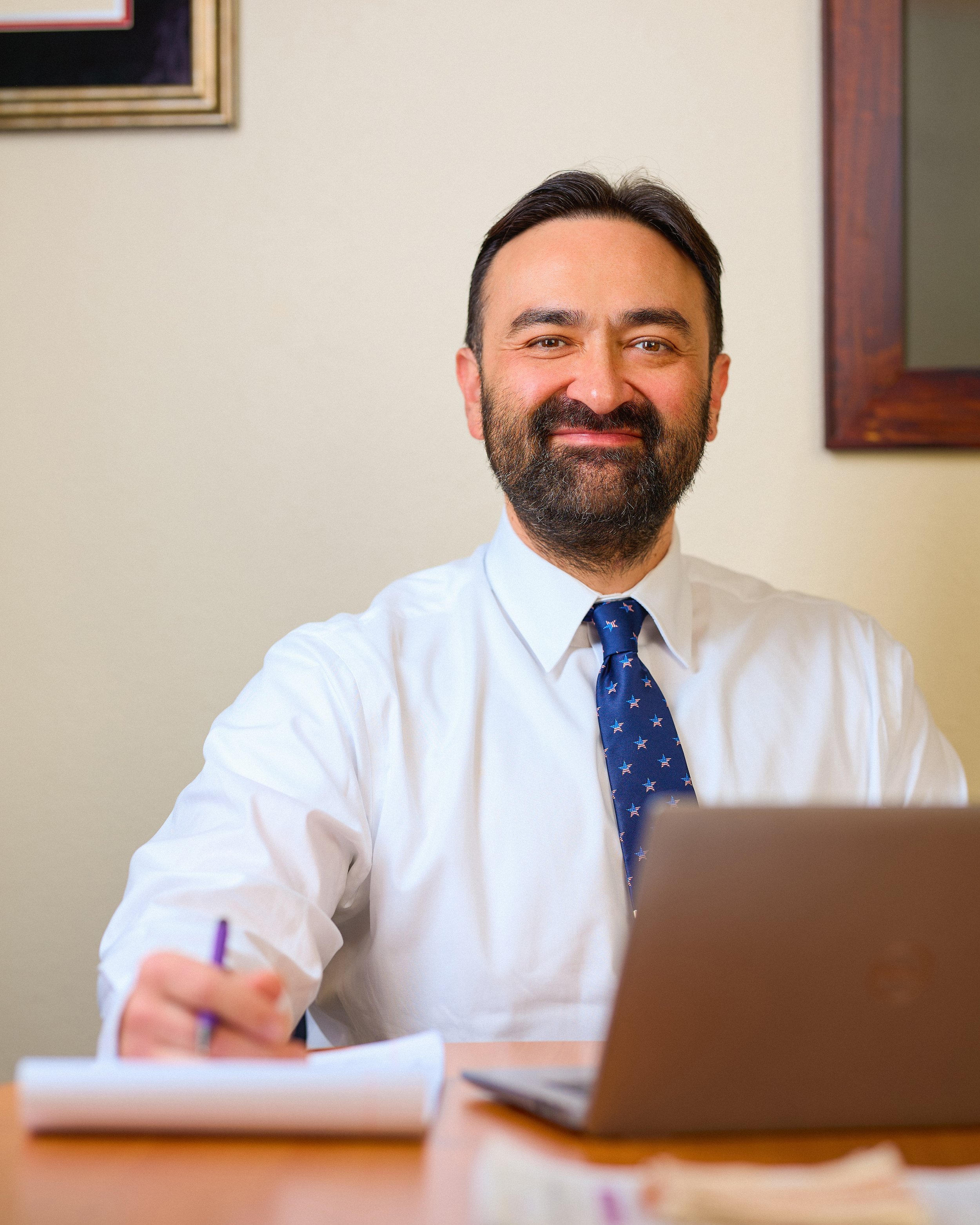 A man in formal attire, with a beard and slicked-back dark hair, smiling while sitting at a desk with a laptop in front of him. He is wearing a white shirt and a patterned tie, writing with a pen.