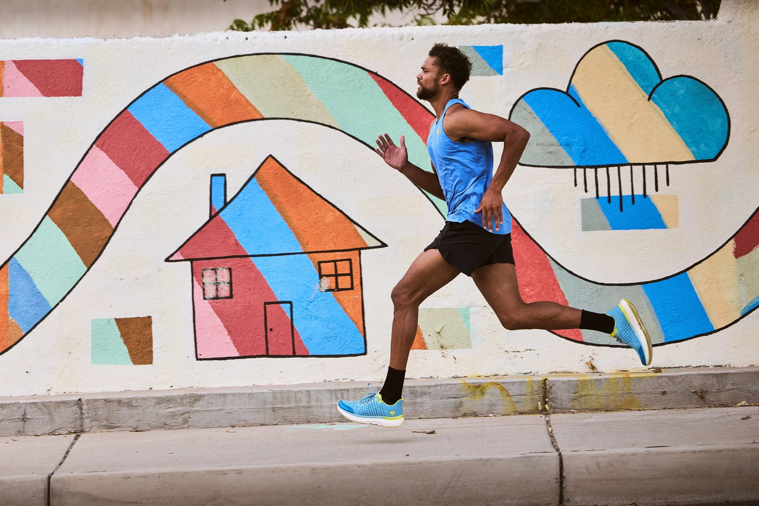 A man running past a colorful mural painted on a wall, featuring abstract shapes, houses, and a rainbow with stripes of a rainbow pattern.