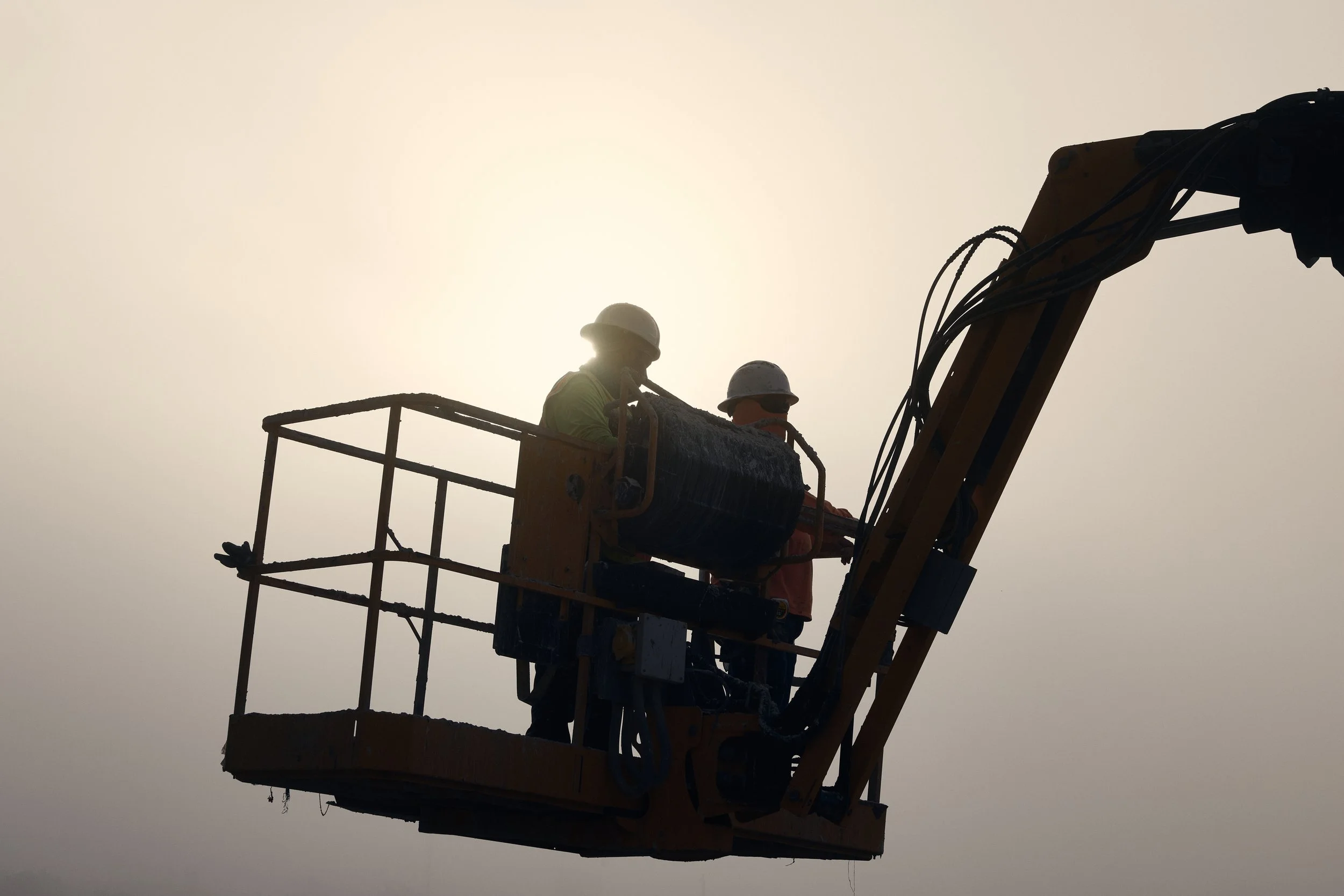 Two construction workers wearing helmets working on a construction lift against a bright, hazy sky.