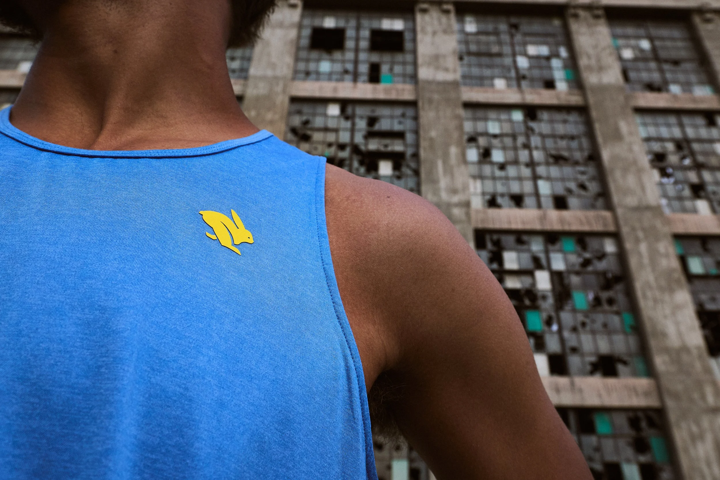 Close-up of a person wearing a bright blue sleeveless top with a yellow rabbit pin, standing in front of an old, dilapidated building with multiple broken and boarded-up windows.